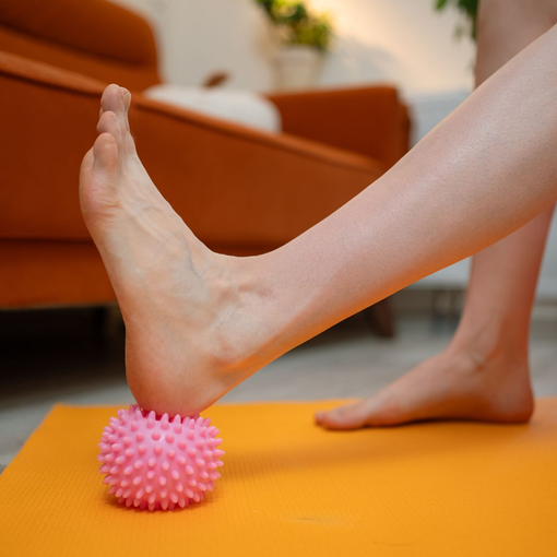 Foot pressing a pink massage ball on an orange yoga mat in a living room