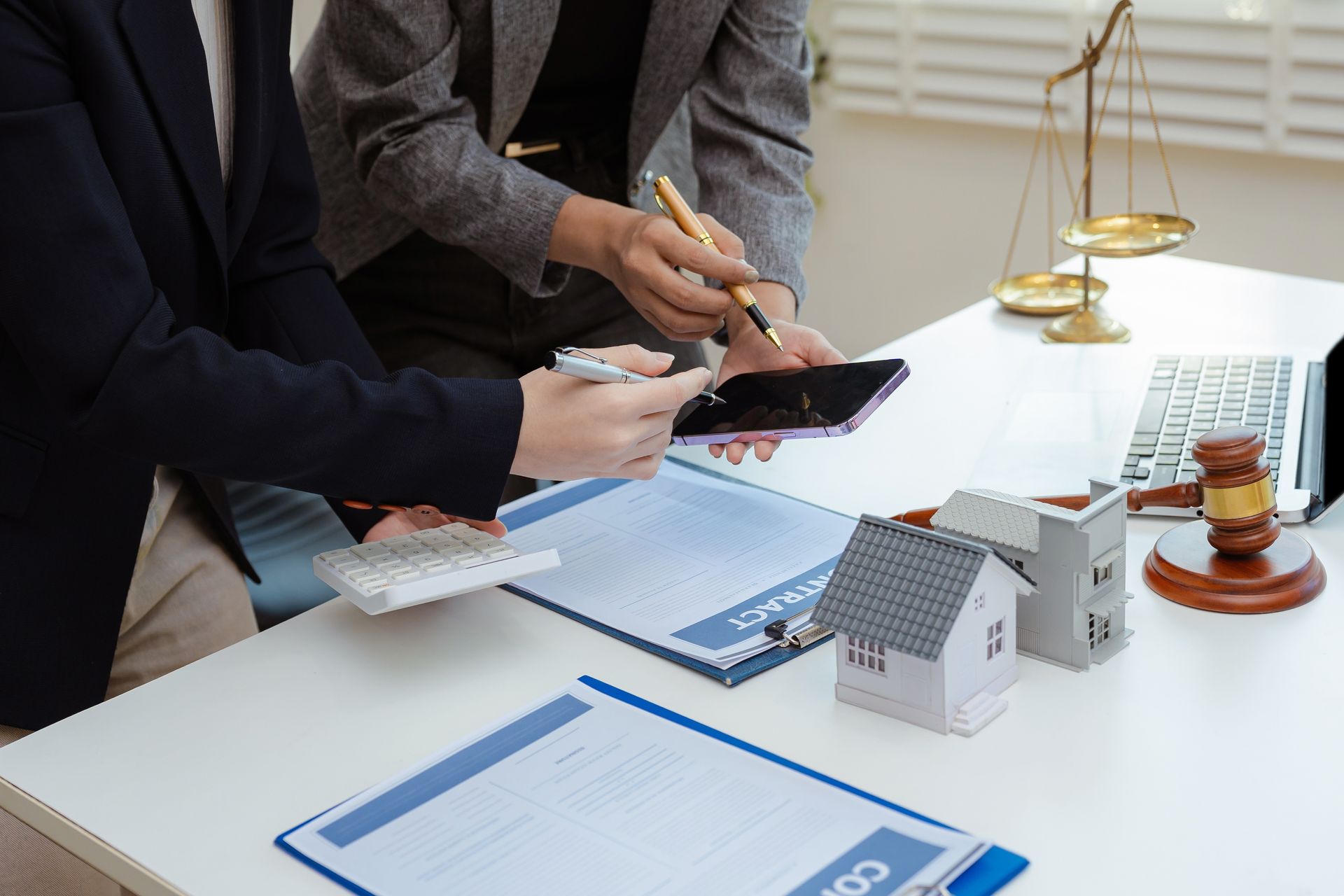 Hands reviewing documents and a tablet at a desk with a house model and scales of justice
