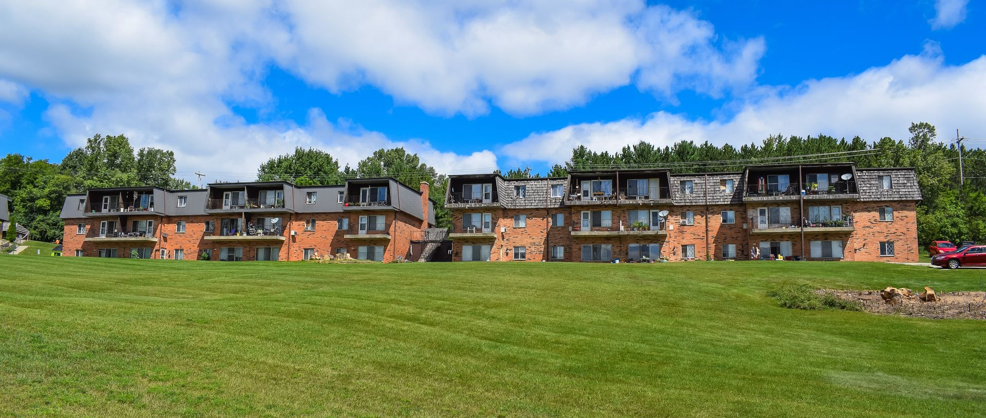 A long, three-story brick apartment building with balconies, surrounded by a green lawn under a bright, cloudy blue sky.