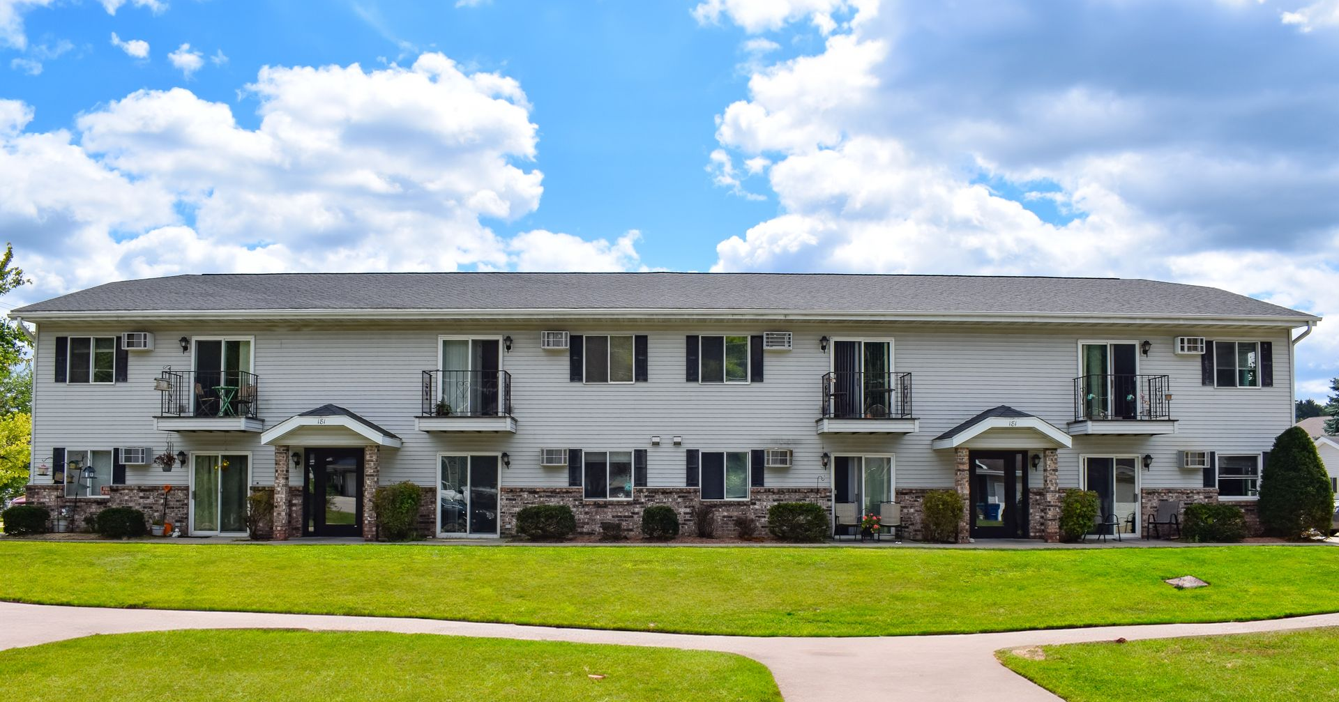 A two-story apartment building with white siding, stone trim, and balconies under a blue sky with scattered clouds.