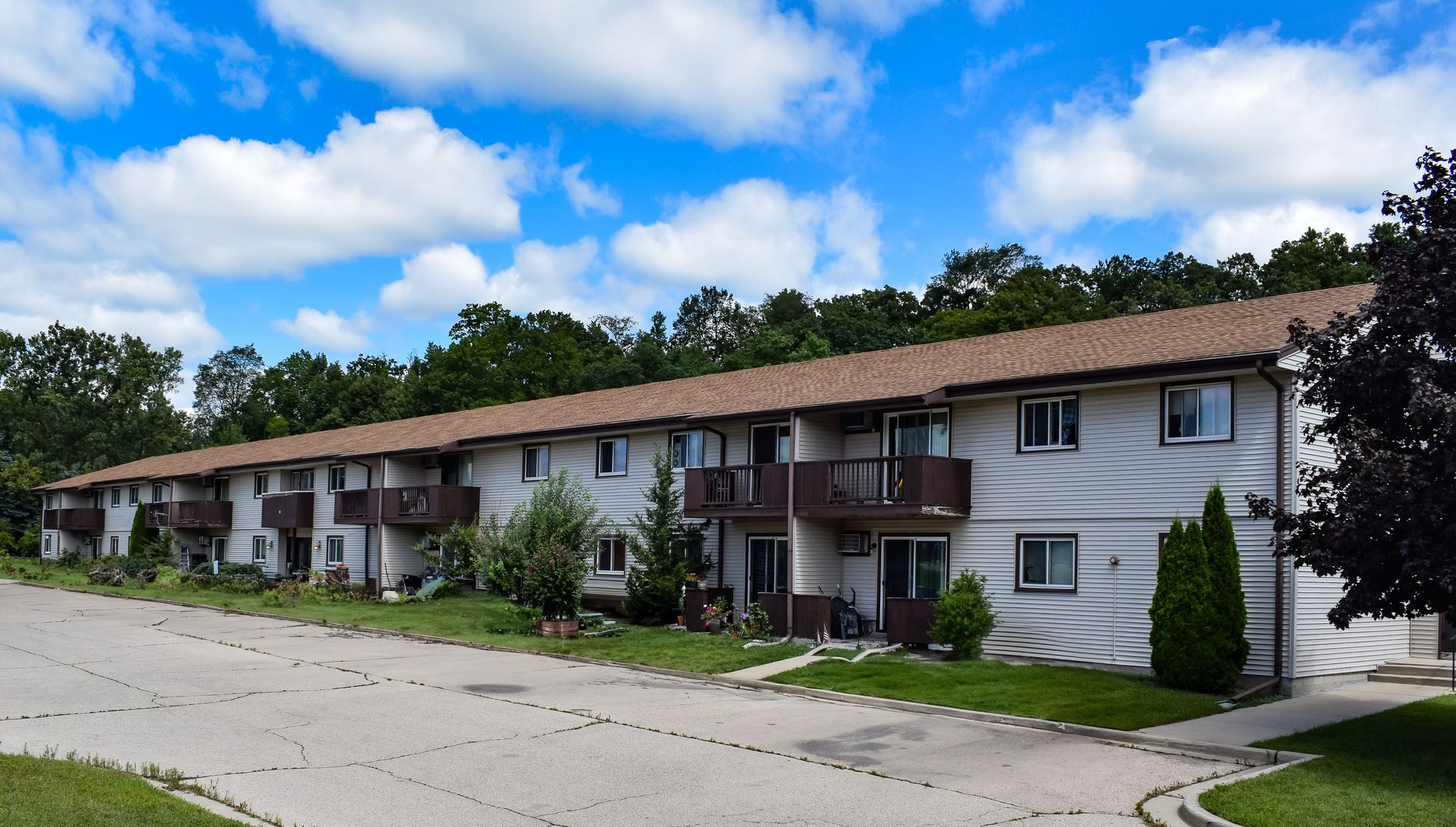 A long, two-story apartment building with white siding, a brown roof, and small balconies under a blue, cloudy sky.