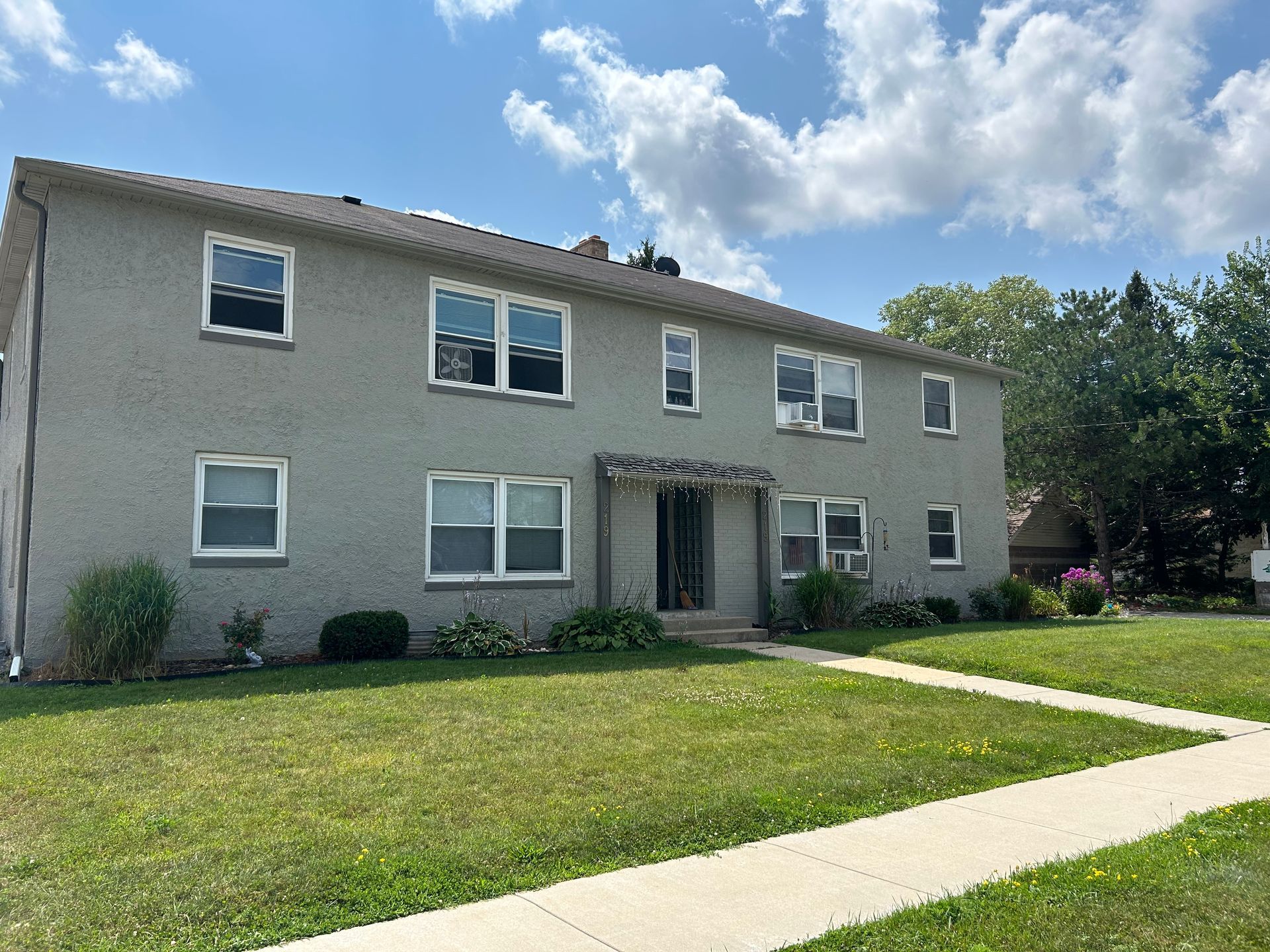 A two-story, gray apartment building with a flat roof and green lawn under a blue, cloudy sky.