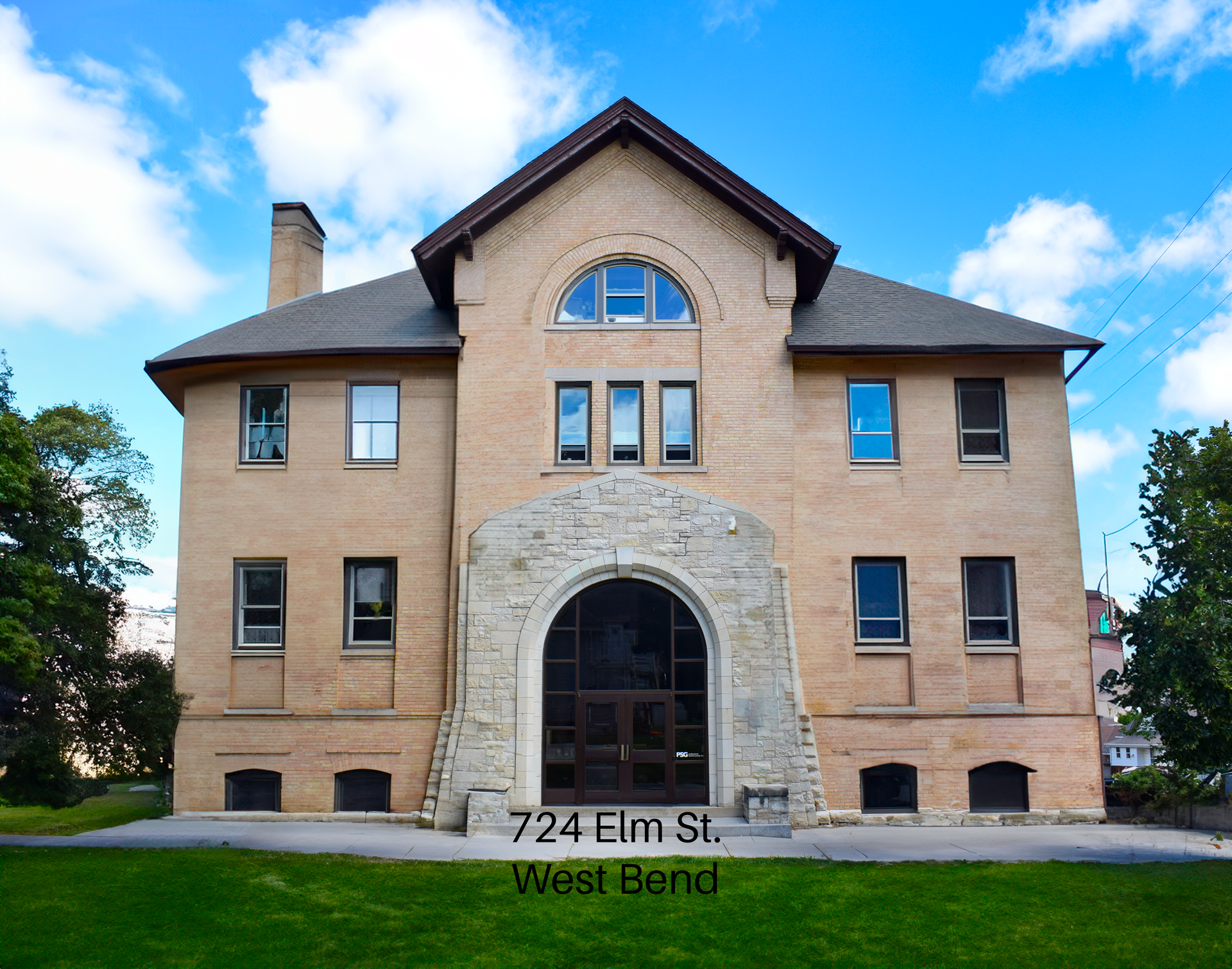 The historic tan brick building at 724 Elm St in West Bend, featuring a stone arched entrance and a semi-circular window.