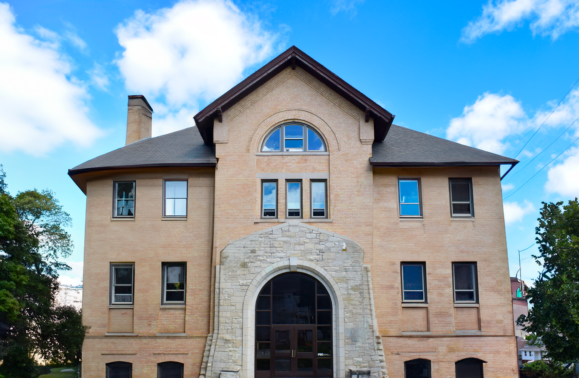 A light tan, two-story brick building with a stone arched entryway and a prominent triangular gable under a blue sky.