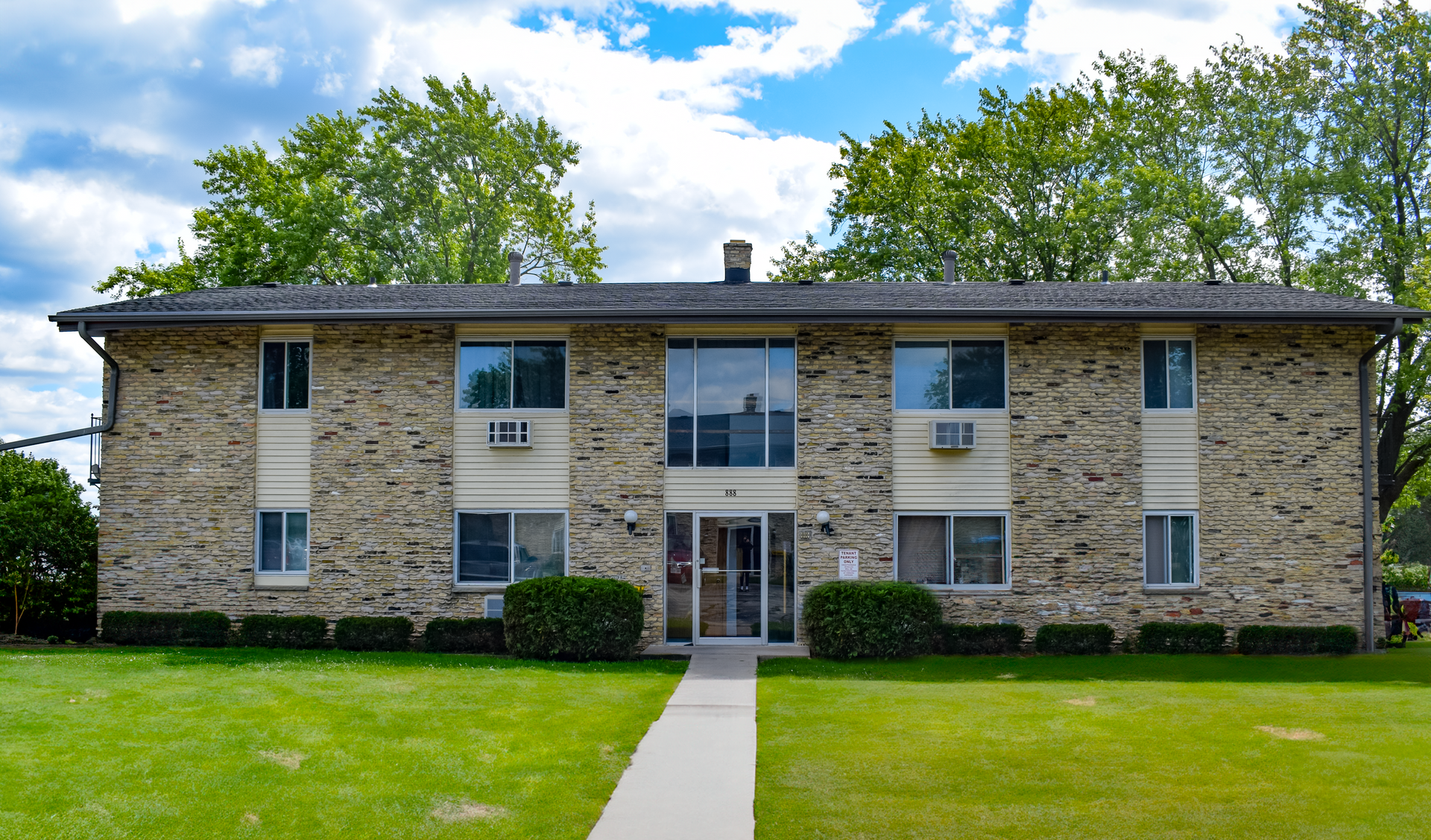 A two-story stone apartment building with a central entrance and walkway, surrounded by a lawn and green trees.