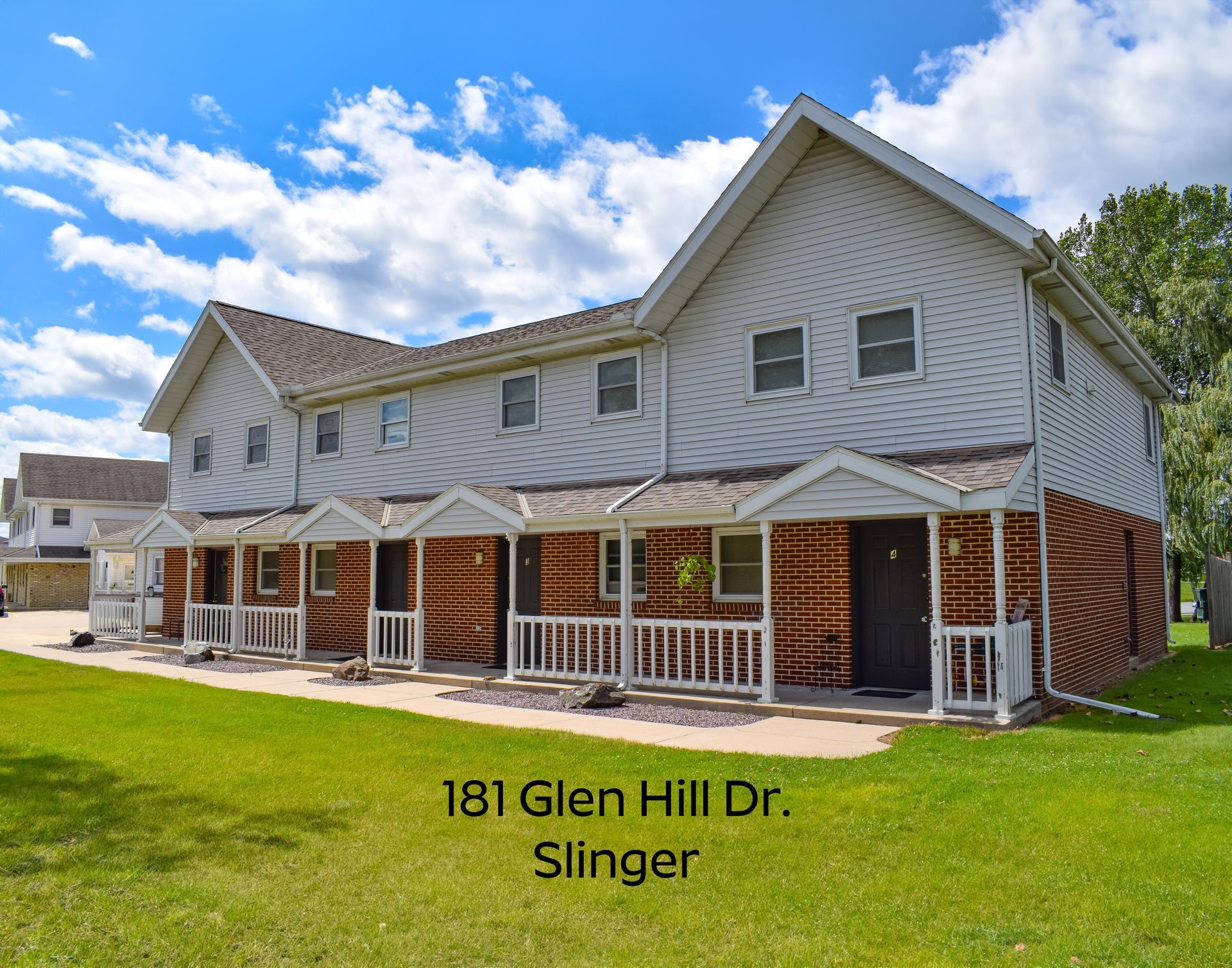 A multi-unit townhome building with light siding and brick accents, featuring small porches, on a grassy lot in Slinger.