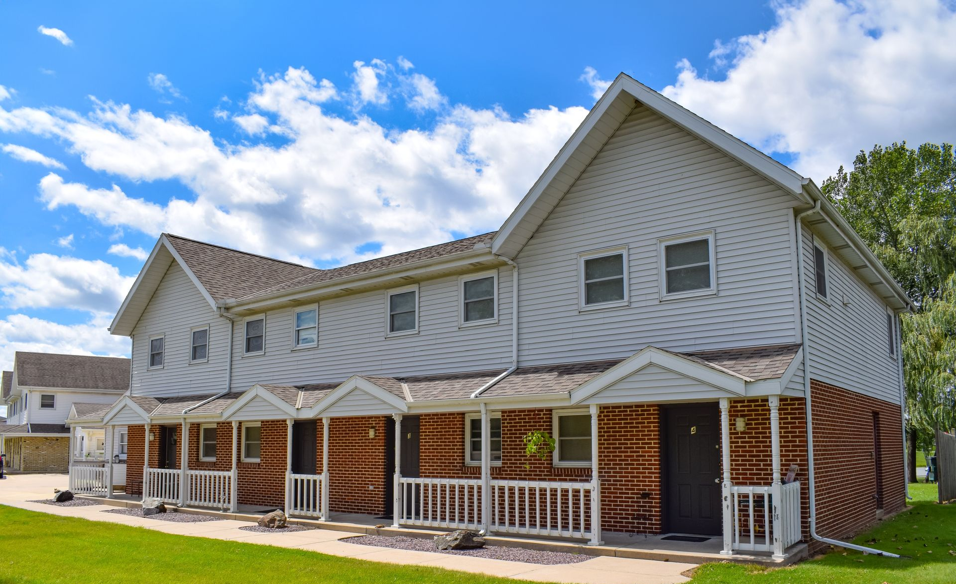 A multi-unit, two-story apartment building with light siding, a brick facade, and individual front porches under a blue sky.