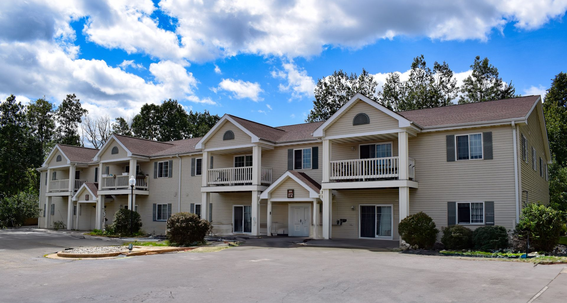 A beige two-story apartment complex with balconies, gabled roofs, and a paved parking lot under a cloudy blue sky.