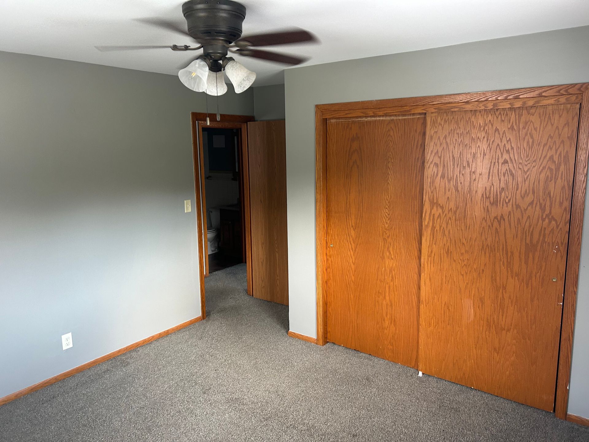 A bedroom with gray walls, carpeted floors, a ceiling fan, a doorway, and a large wooden closet.