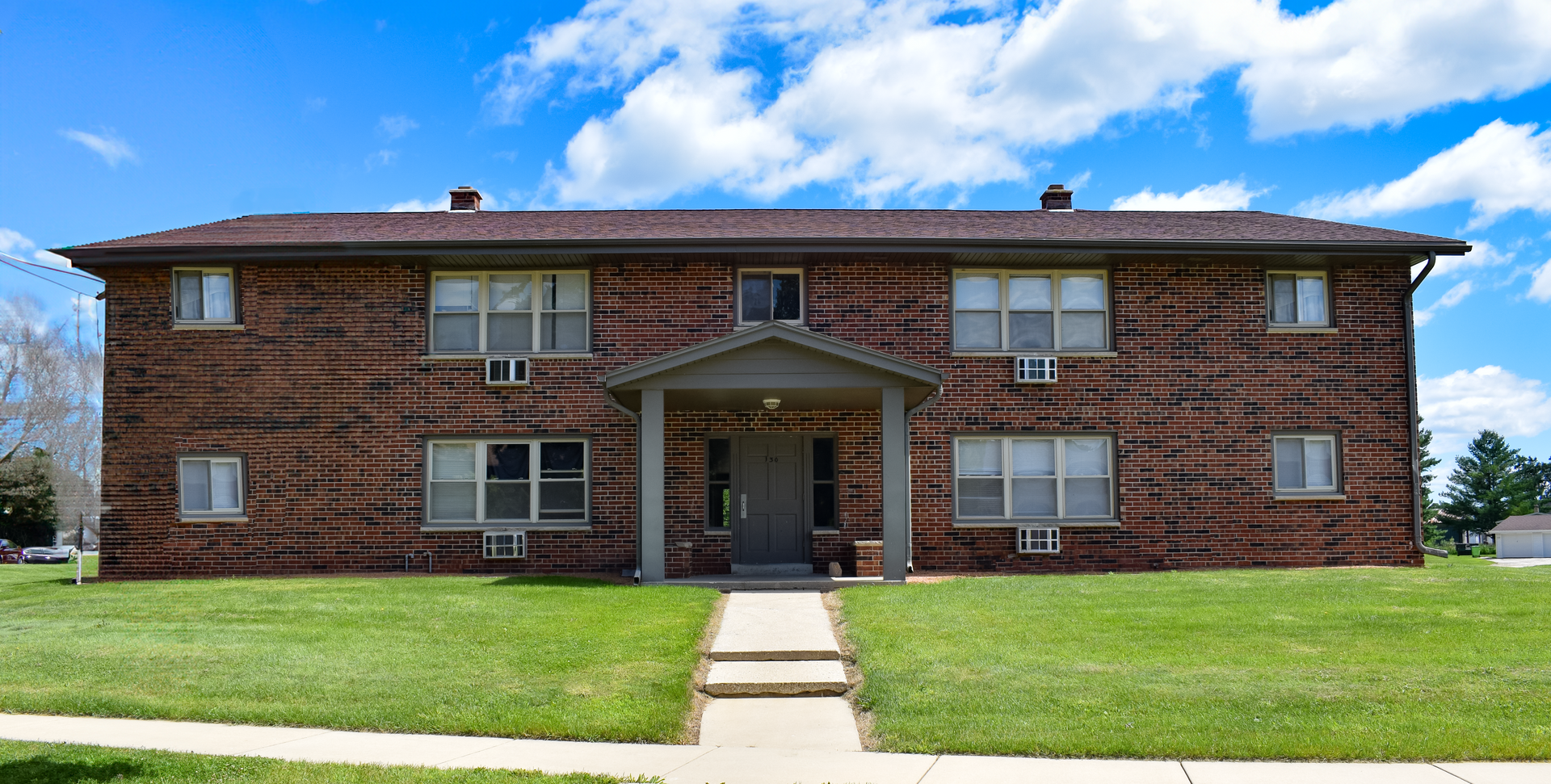 A two-story apartment building with dark horizontal siding, a central entryway, and a grassy front lawn under a blue sky.