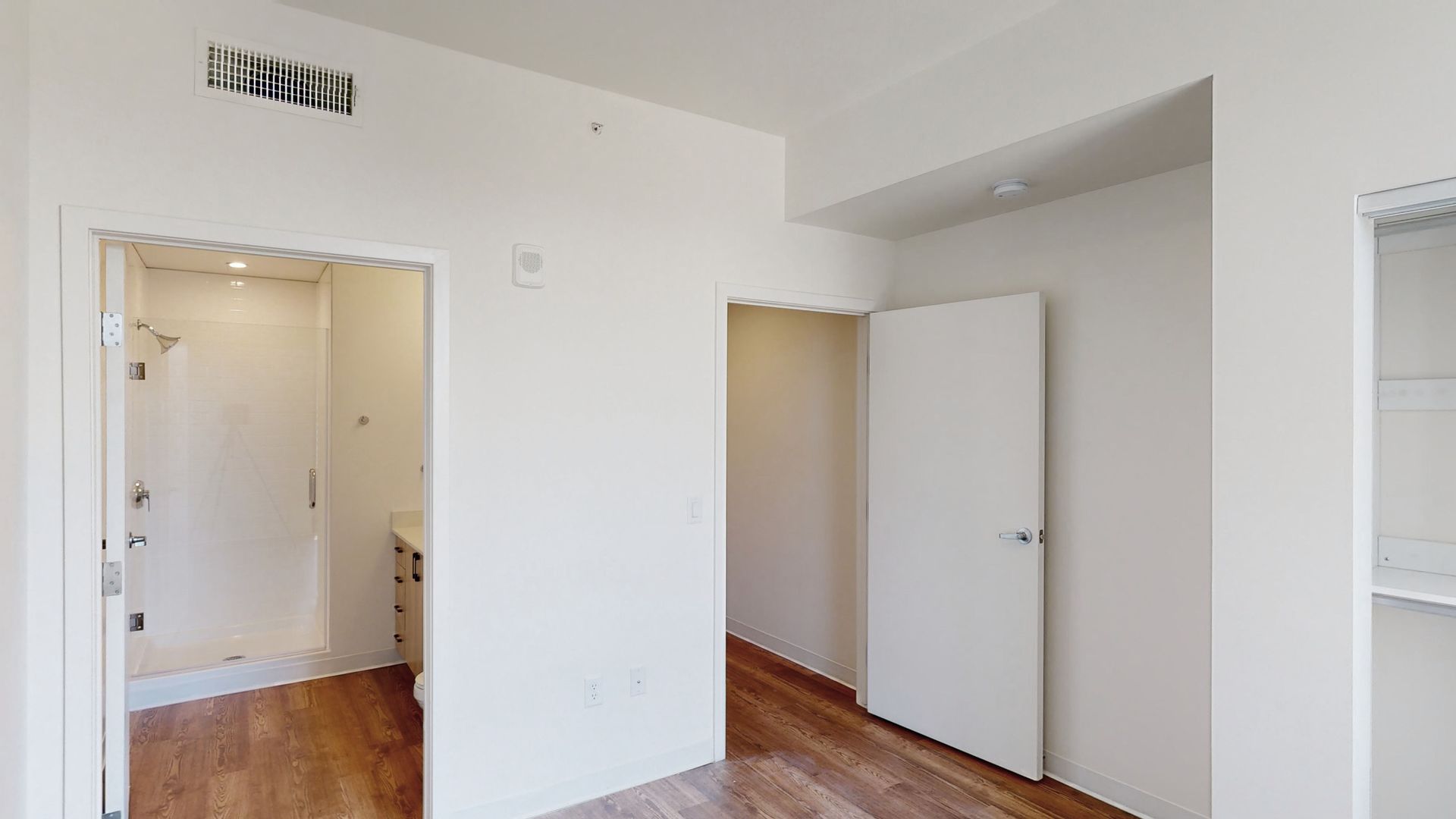 A bedroom with hardwood floors , white walls and a door leading to a bathroom.