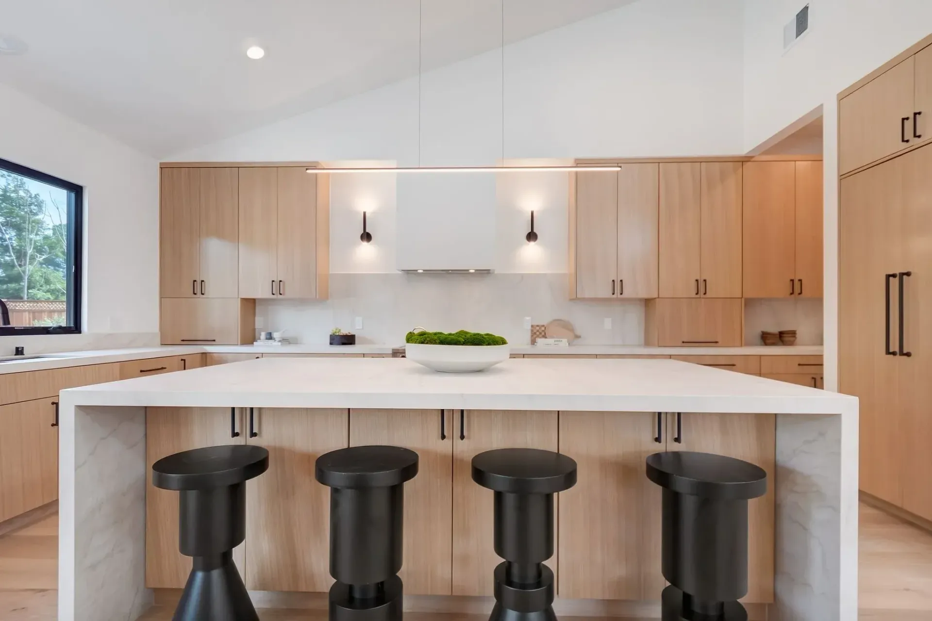 Modern kitchen with light wood cabinets, white countertops, and black stools.