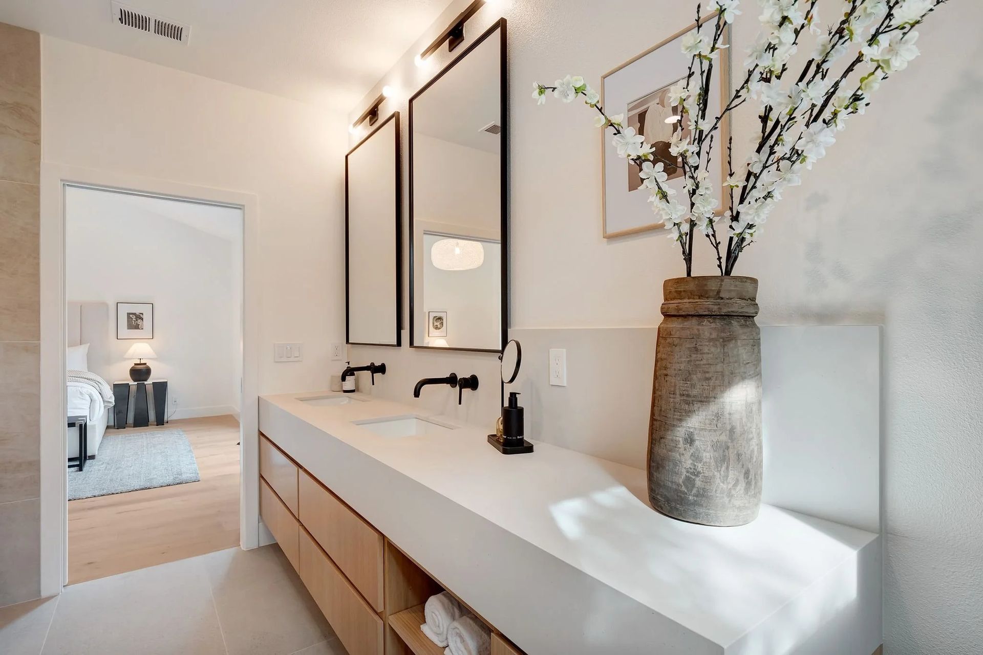 Modern bathroom with double sinks, mirrors, and a vase of flowers on a white countertop; view into a bedroom.