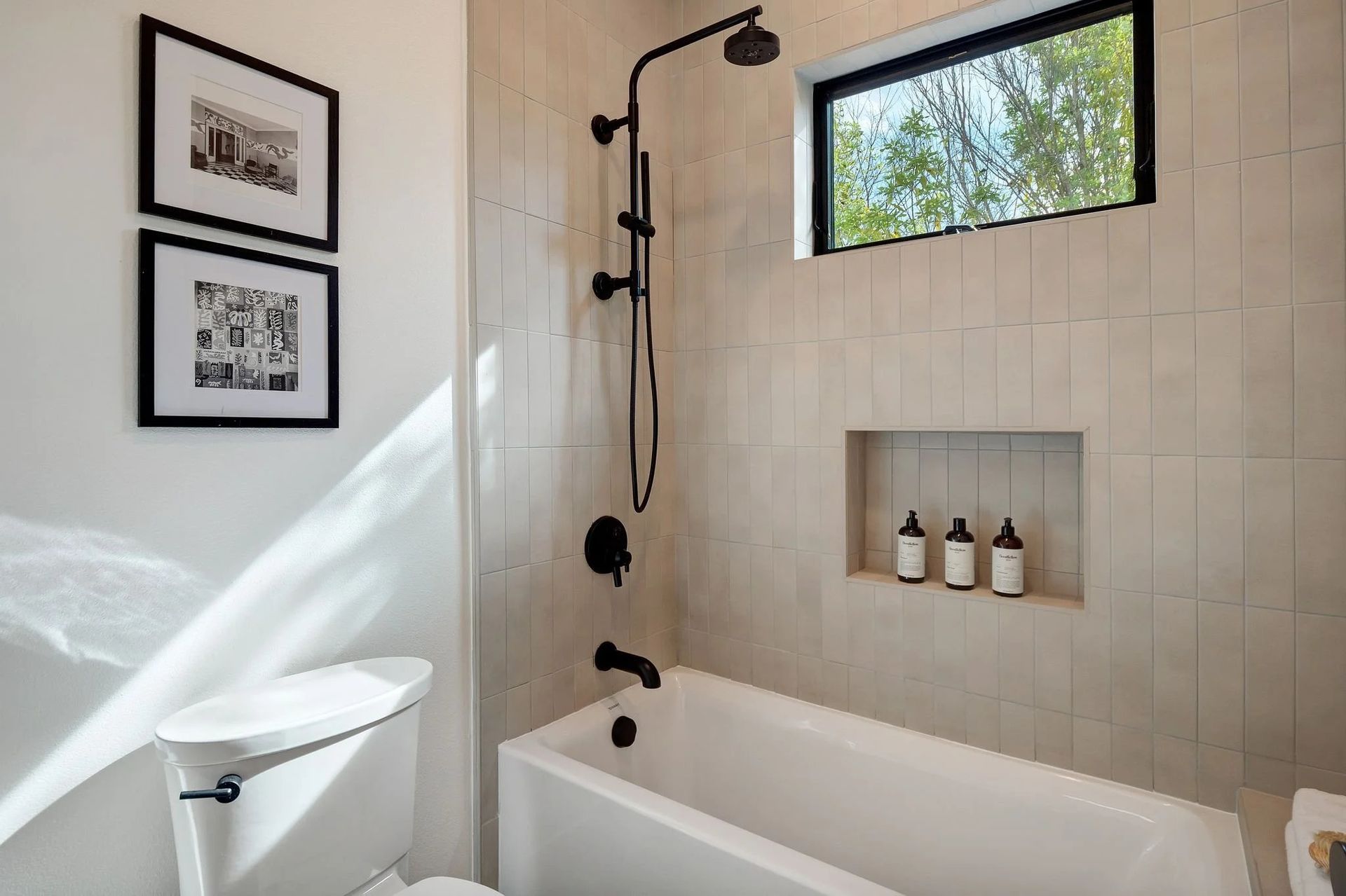 Bathroom with white tub, shower, toilet, and black fixtures; window, built-in shelf, art on wall.