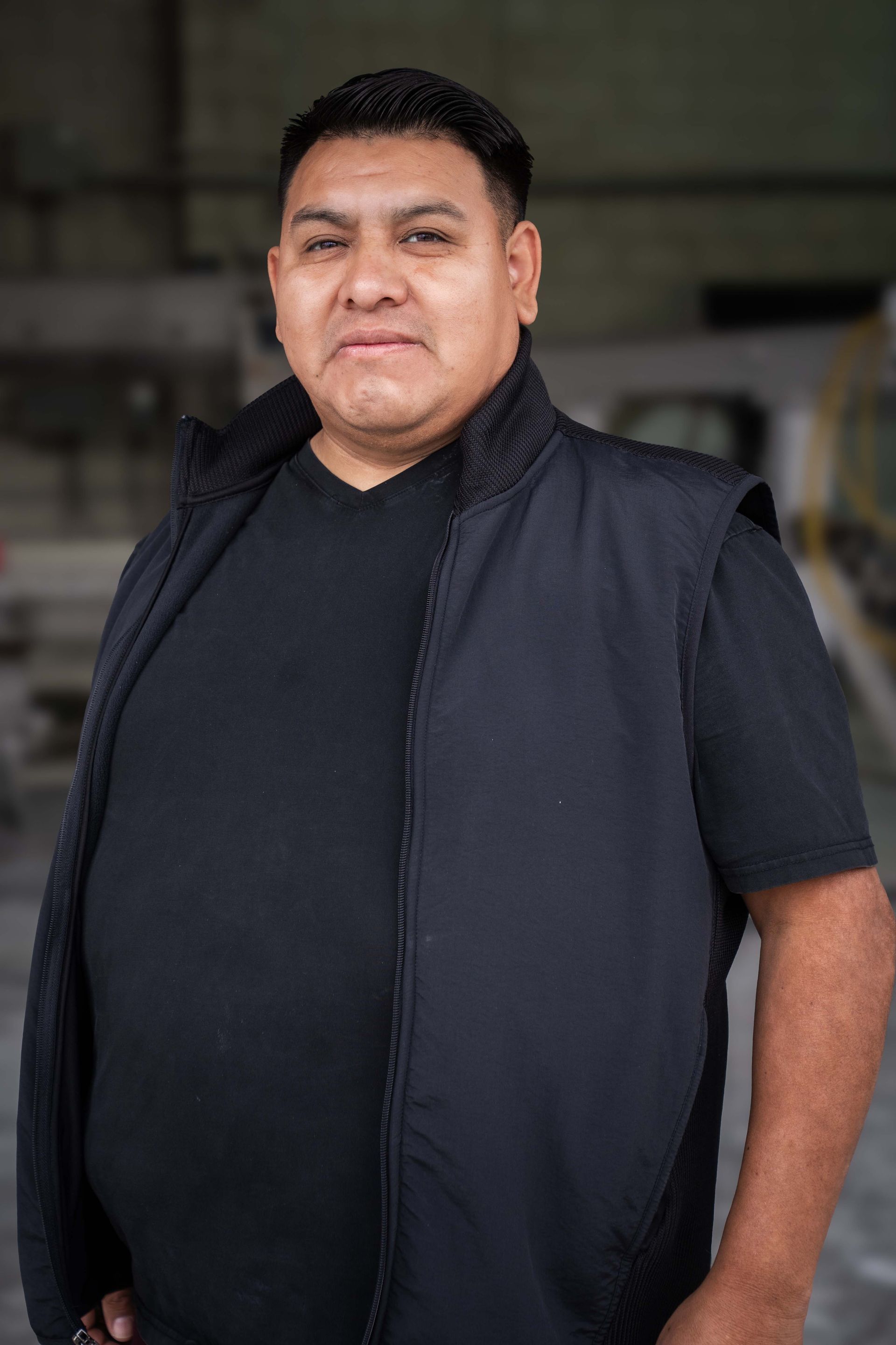Man wearing black vest and shirt in an industrial setting, looking forward.