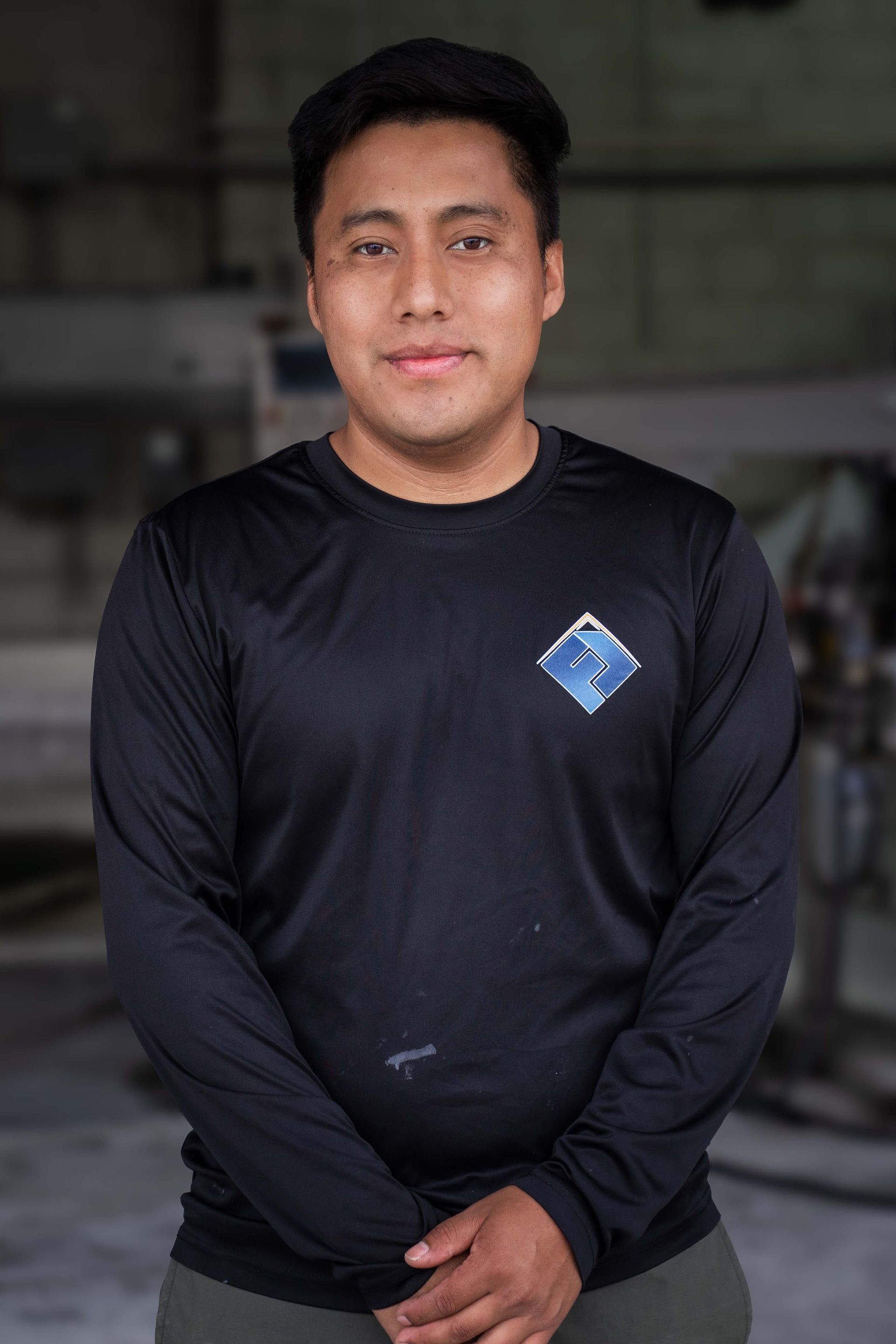 Man in black long-sleeve shirt with logo, smiling, in industrial setting.