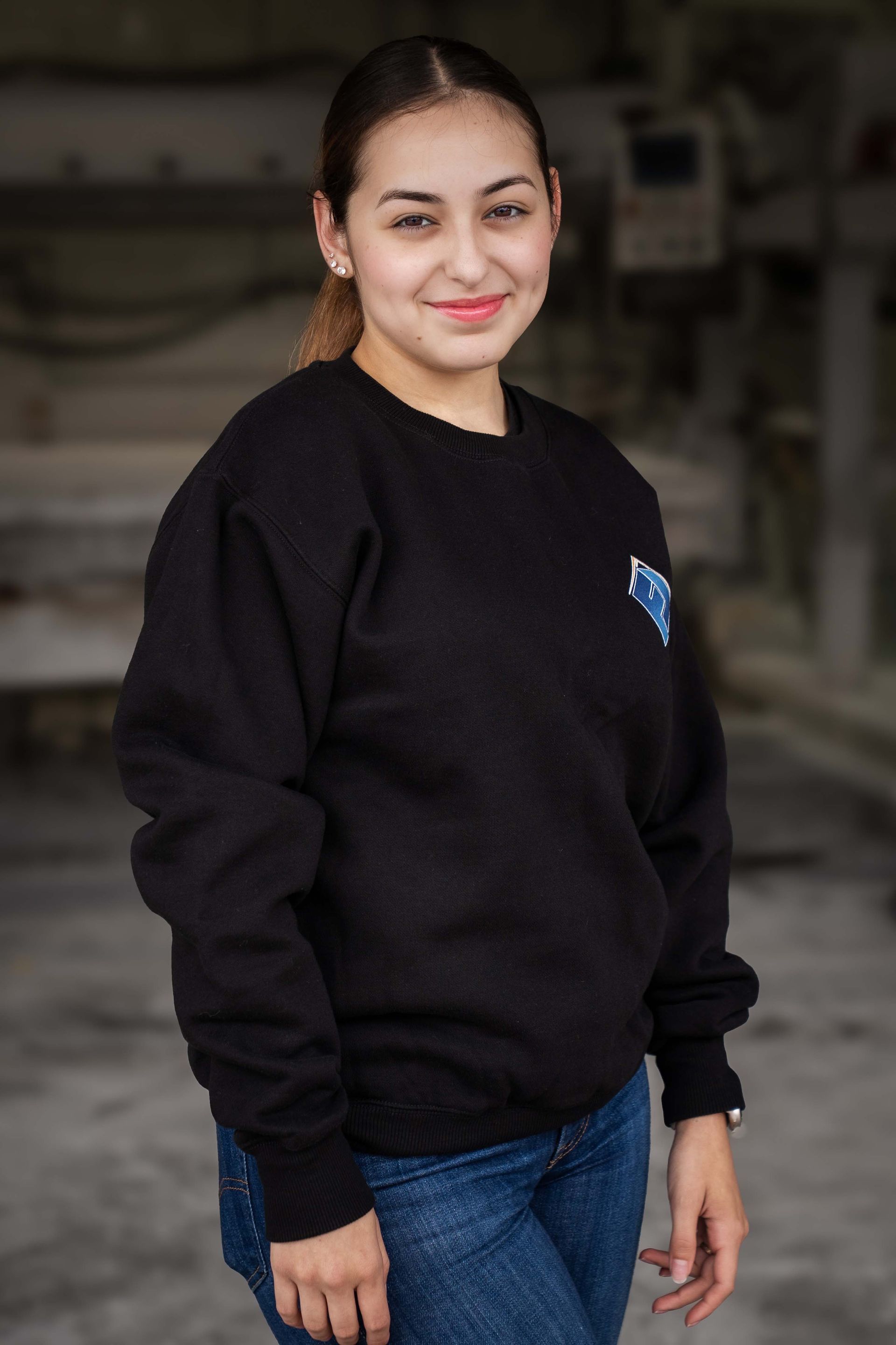 Woman in a black sweatshirt and jeans smiles in an industrial setting.