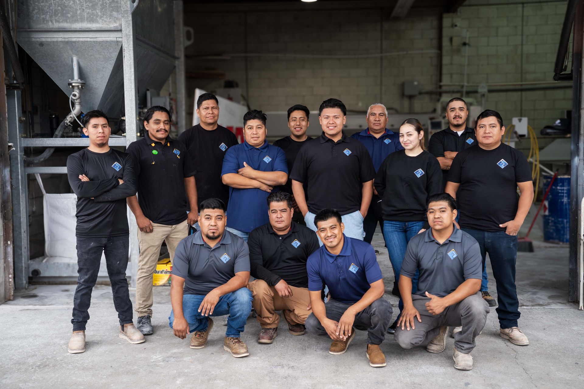 Group of factory workers posing; mostly men wearing black/blue shirts; industrial setting.
