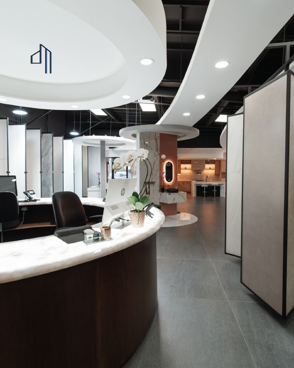 Reception desk in showroom. Curved brown base, light-colored top, computer, and two chairs under a circular recessed ceiling with lights.