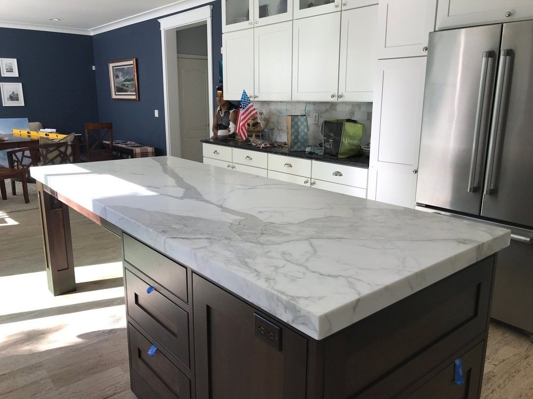 Kitchen with white marble island countertop and dark cabinets, stainless steel refrigerator, blue wall.