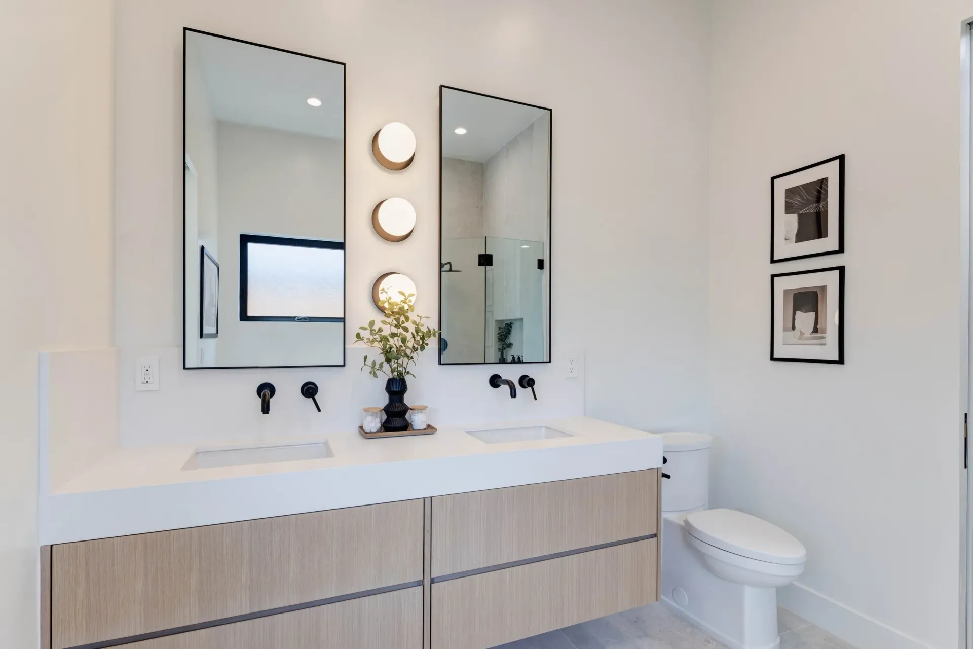 Modern bathroom with double vanity, two mirrors, and a toilet. Bright white walls, wood cabinets.