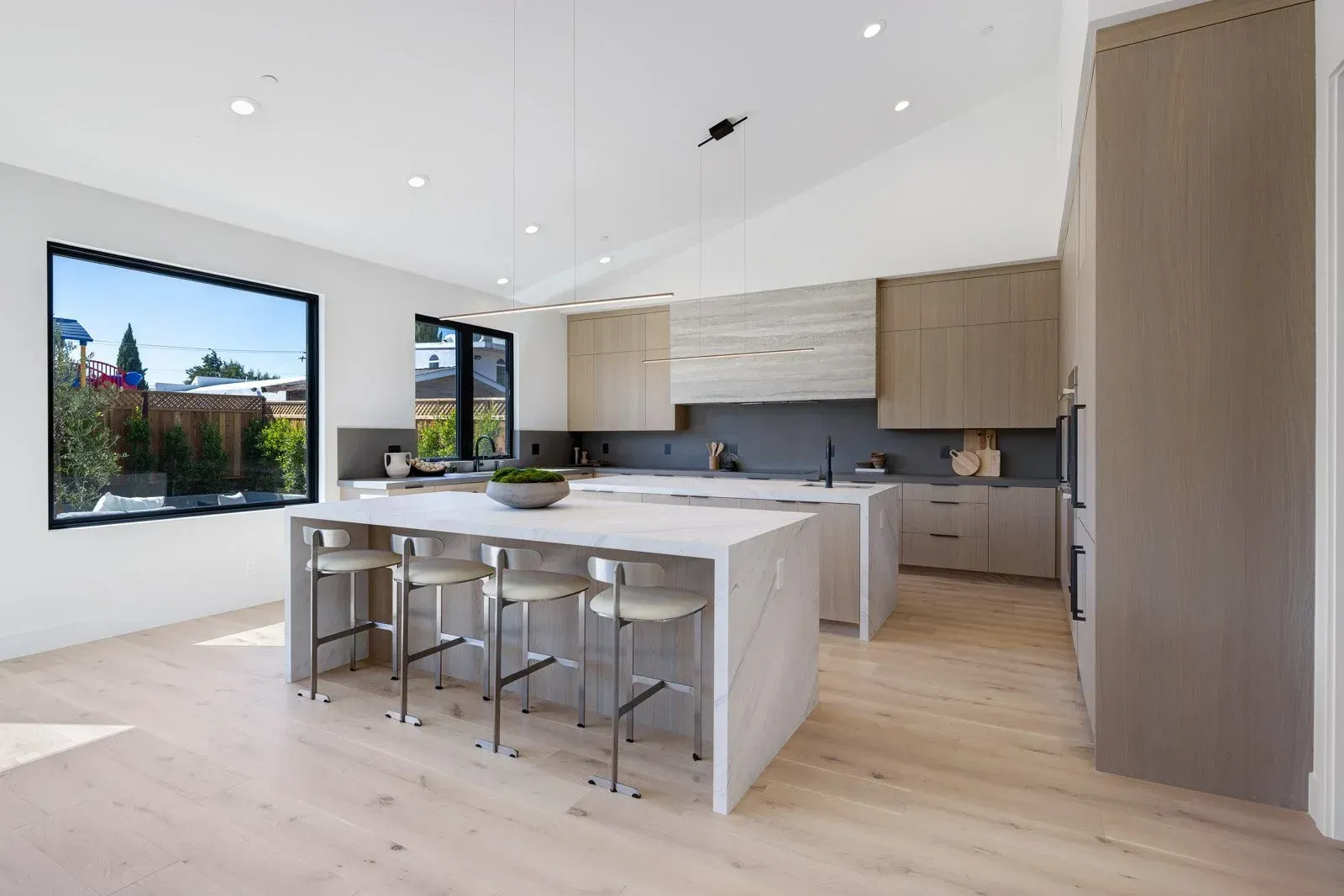 Modern kitchen with a large island, light wood floors, and large windows overlooking greenery.