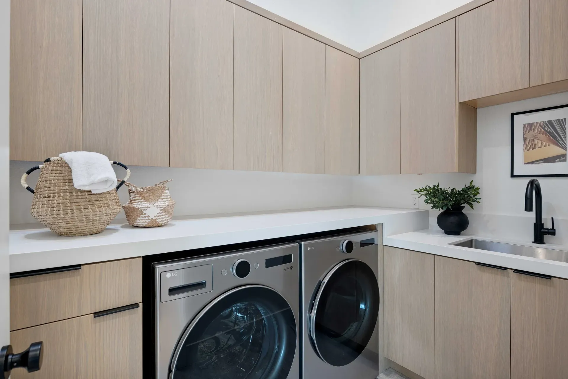 Laundry room with light wood cabinets, stainless steel washer and dryer, and a small sink.