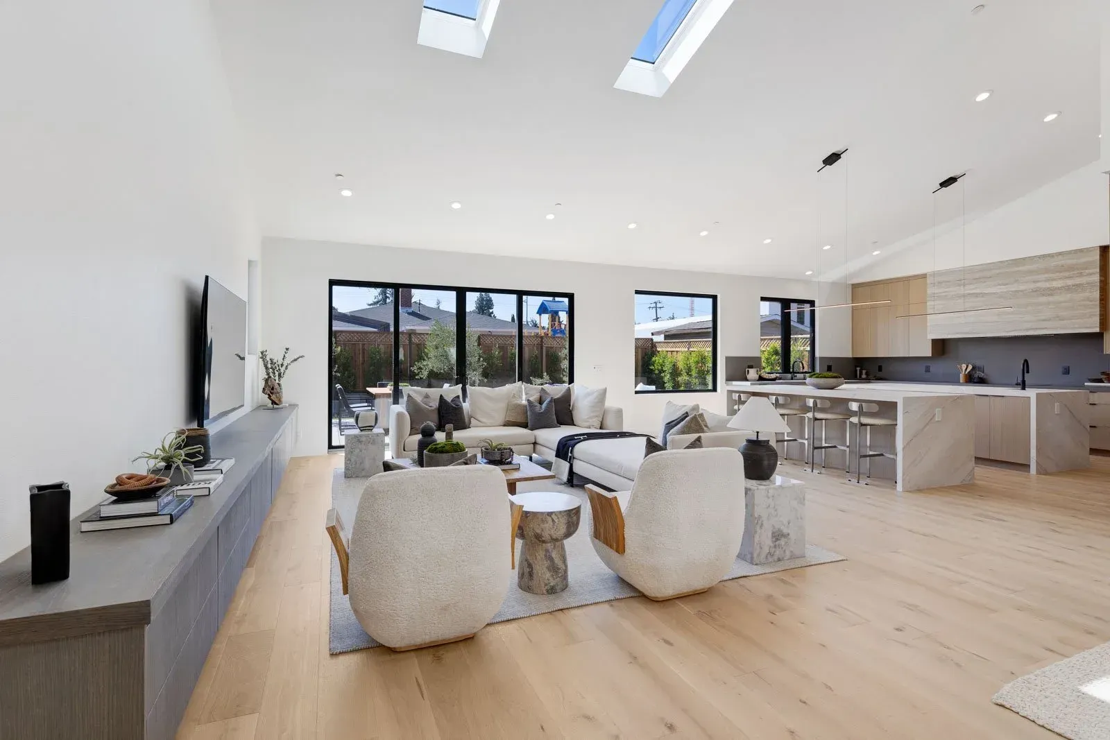 Spacious, modern living room with light wood floors, neutral furniture, and kitchen in background.