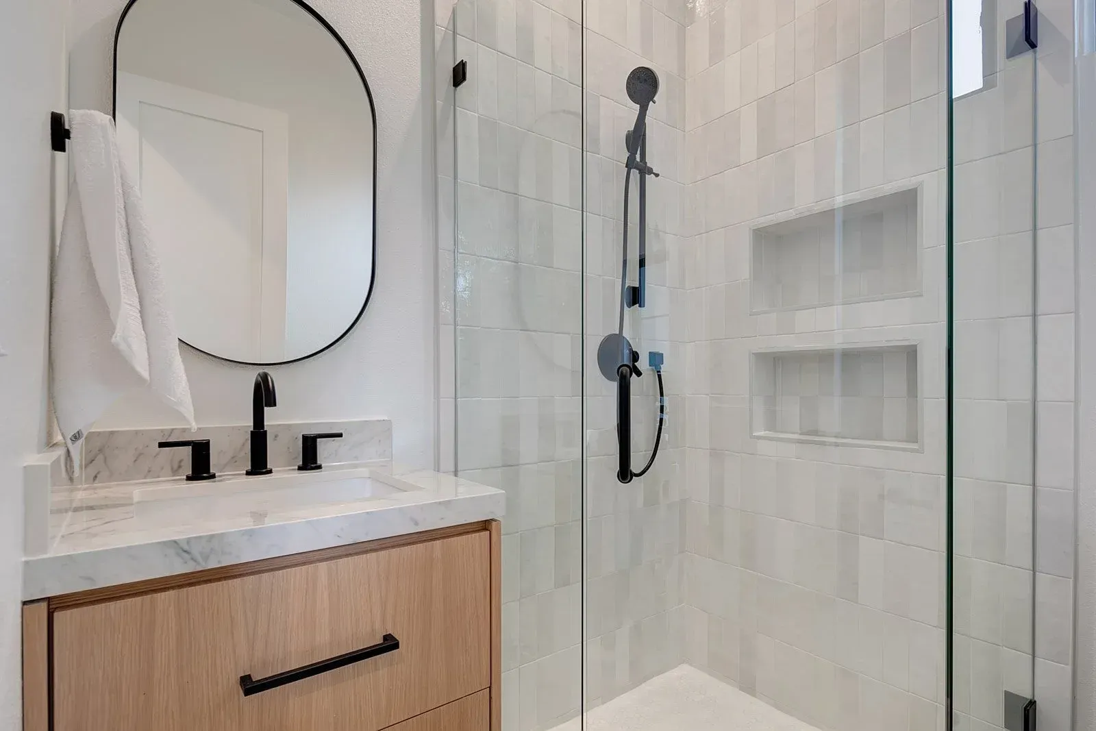 Modern bathroom with wood vanity, oval mirror, and glass shower with white tiled walls.