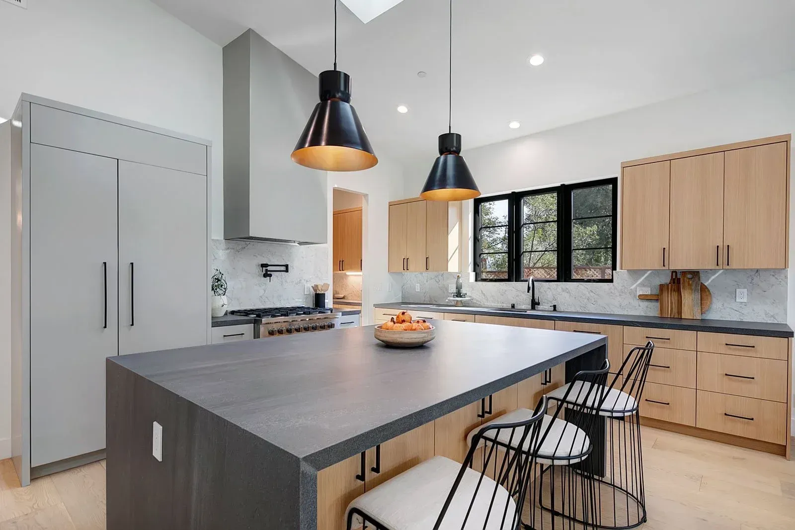 Modern kitchen with gray island, light wood cabinets, black pendant lights, and a window.