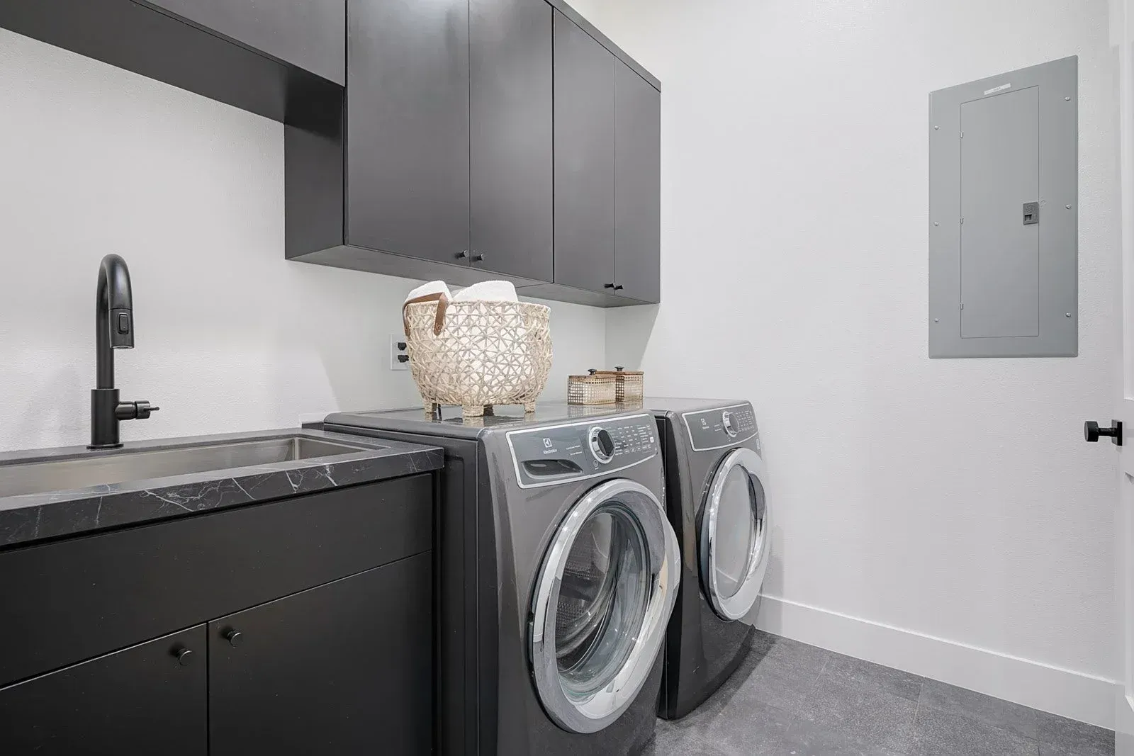 Laundry room with dark gray cabinets, sink, washing machine, and electrical panel on white walls.