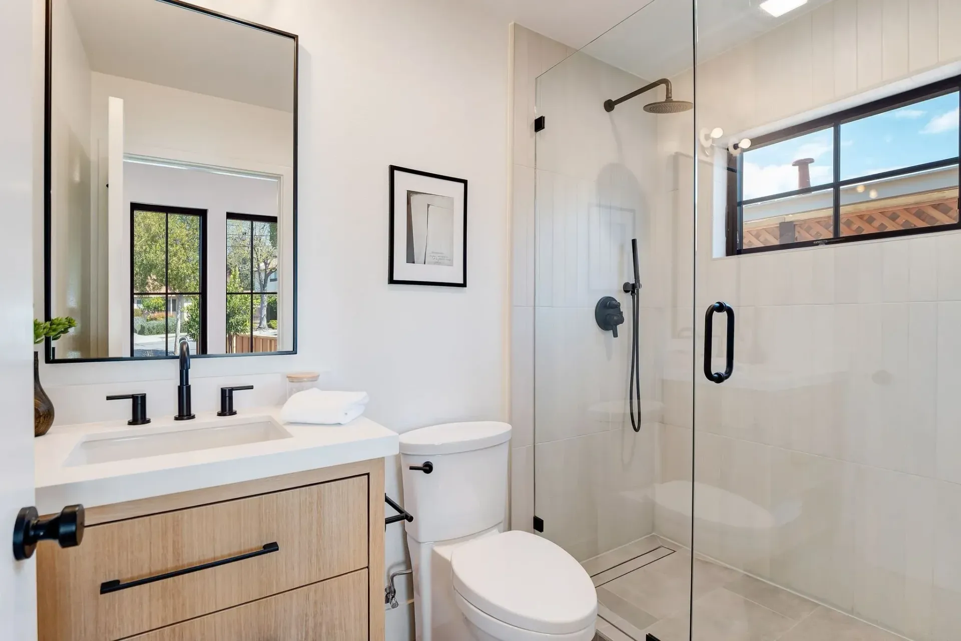 Modern bathroom with white walls, light wood vanity, glass shower, and a window.