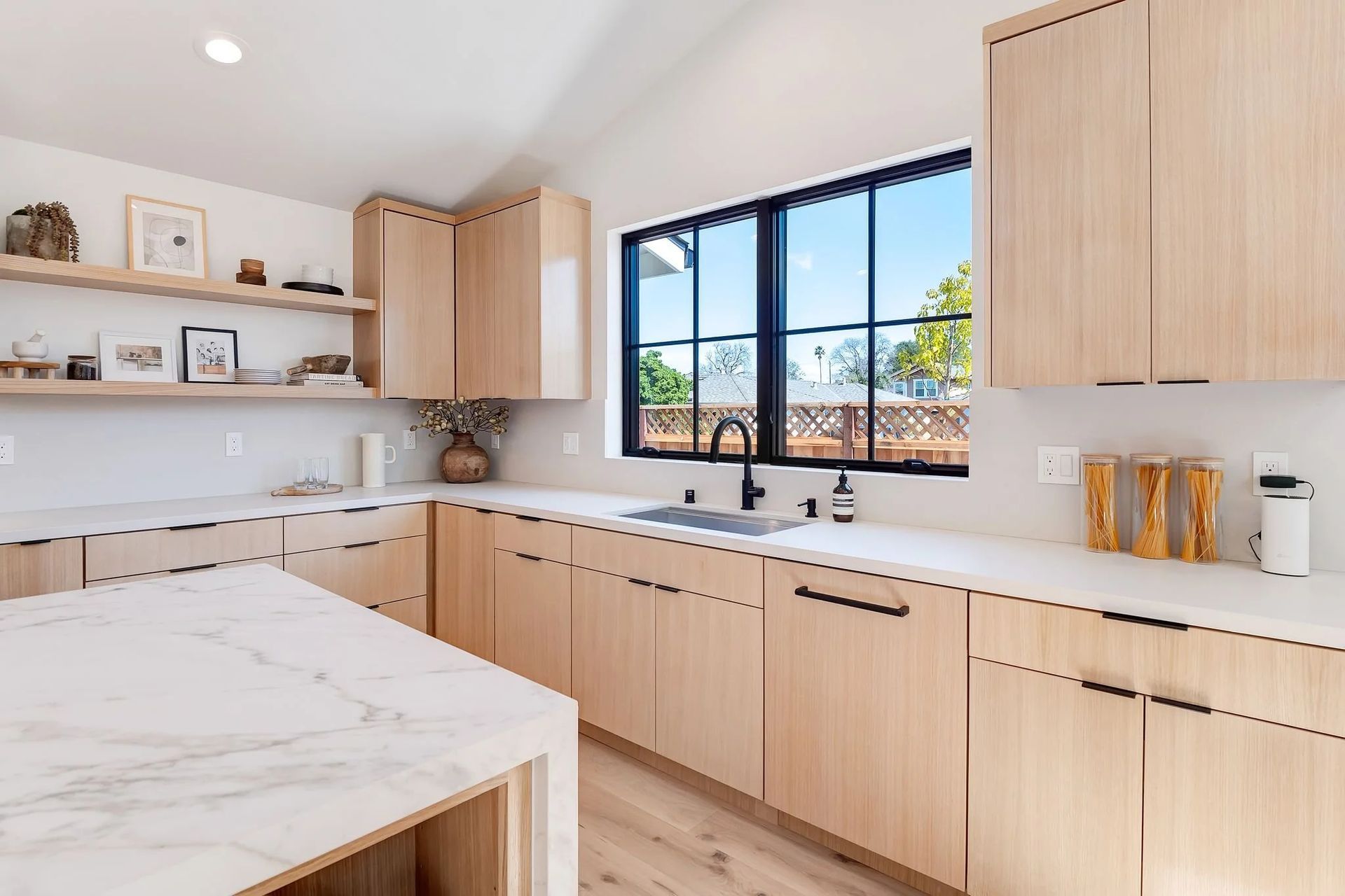 Modern kitchen with light wood cabinets, marble countertops, and a large window overlooking a green landscape.