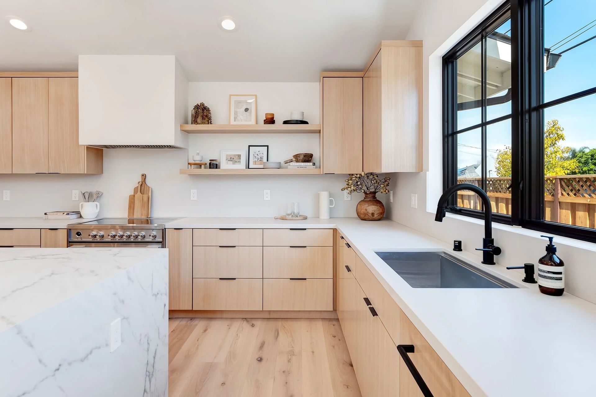 Modern kitchen with light wood cabinets, white countertops, large window.