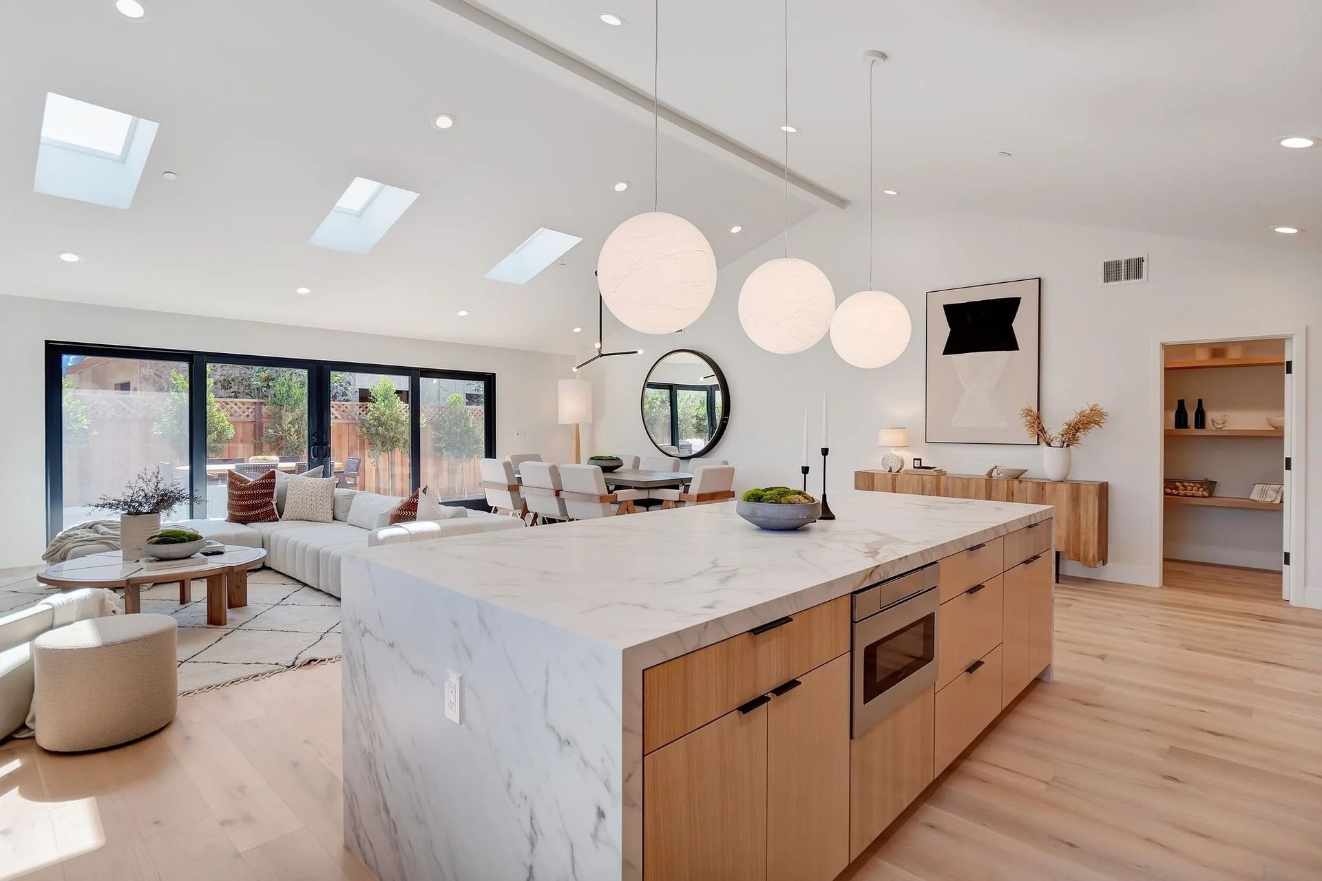 Open-concept kitchen with island, light wood cabinets, marble countertop, and pendant lights, leading to living area.