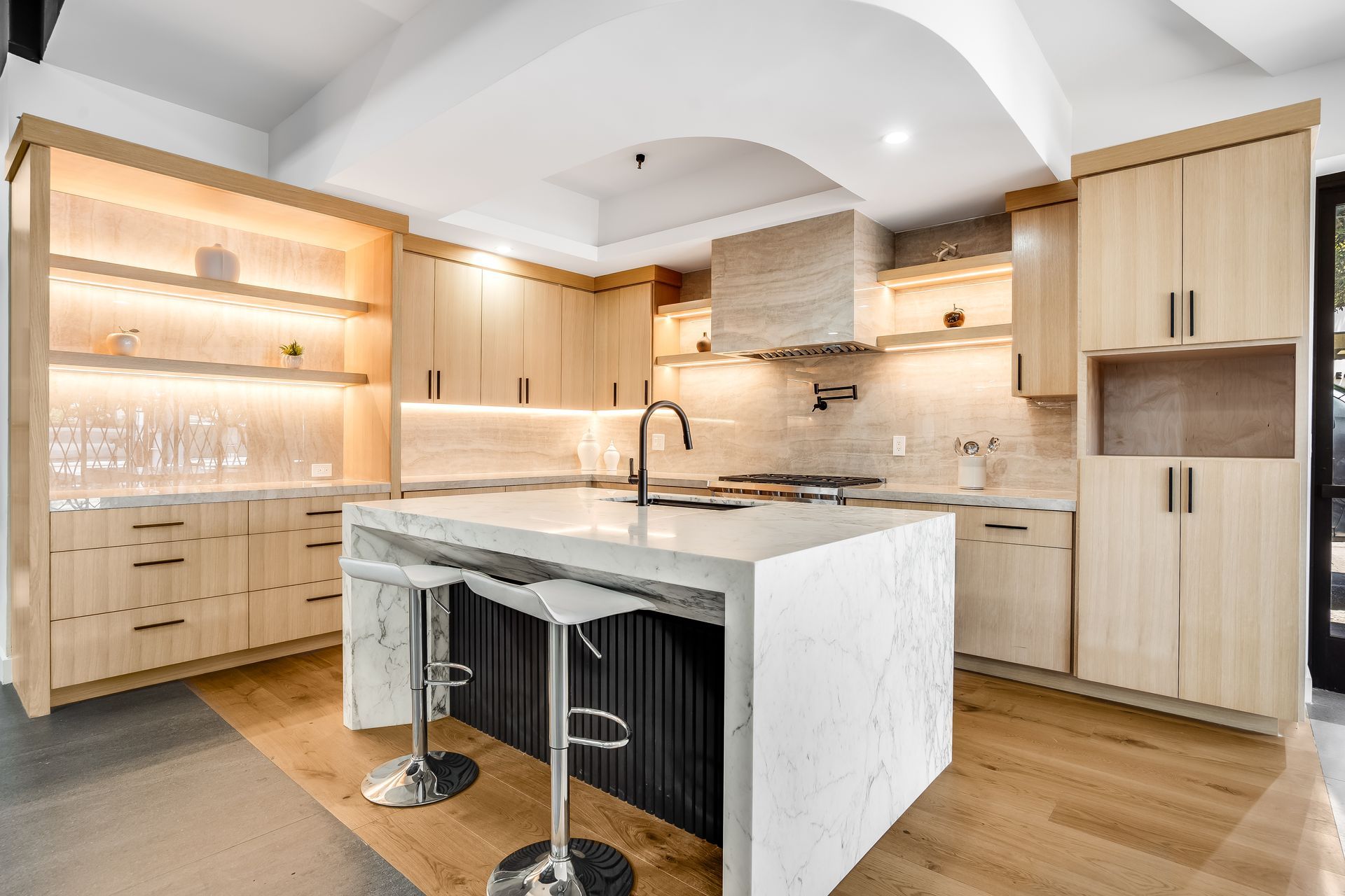 Modern kitchen with light wood cabinets, marble countertops, and island with black base and silver stools.