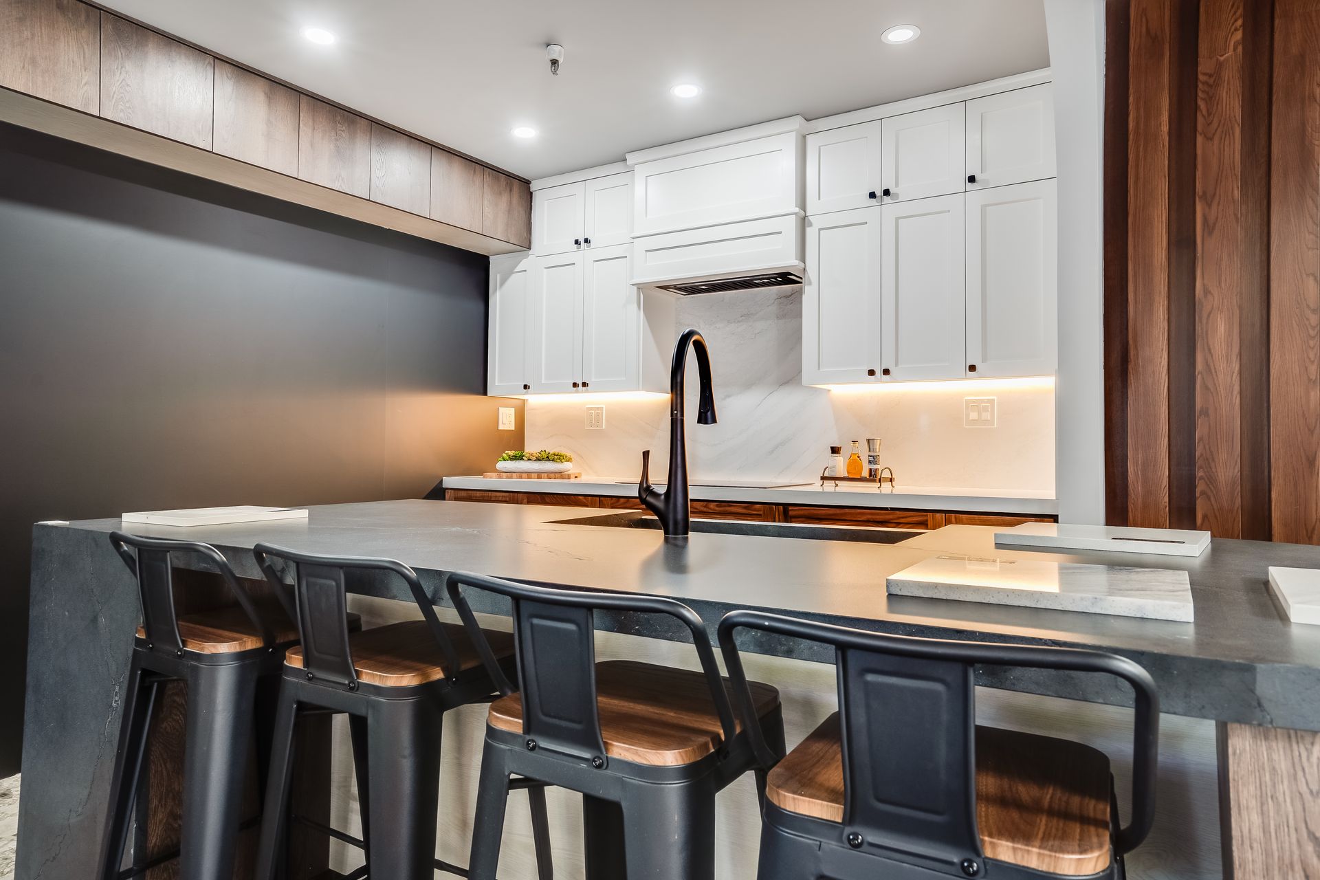 Modern kitchen with white cabinets, dark countertop, black stools, and wooden accents.