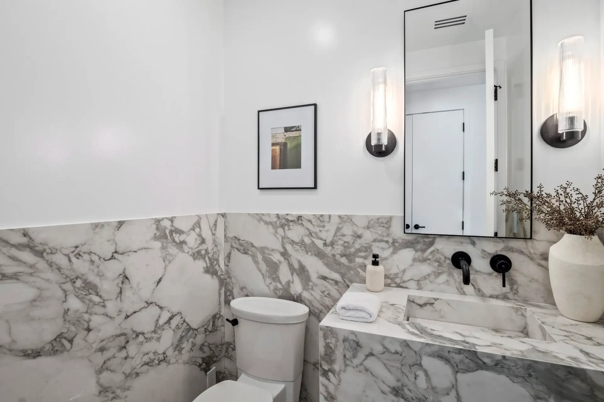 White and marble bathroom with a toilet, sink, mirror, and sconces.