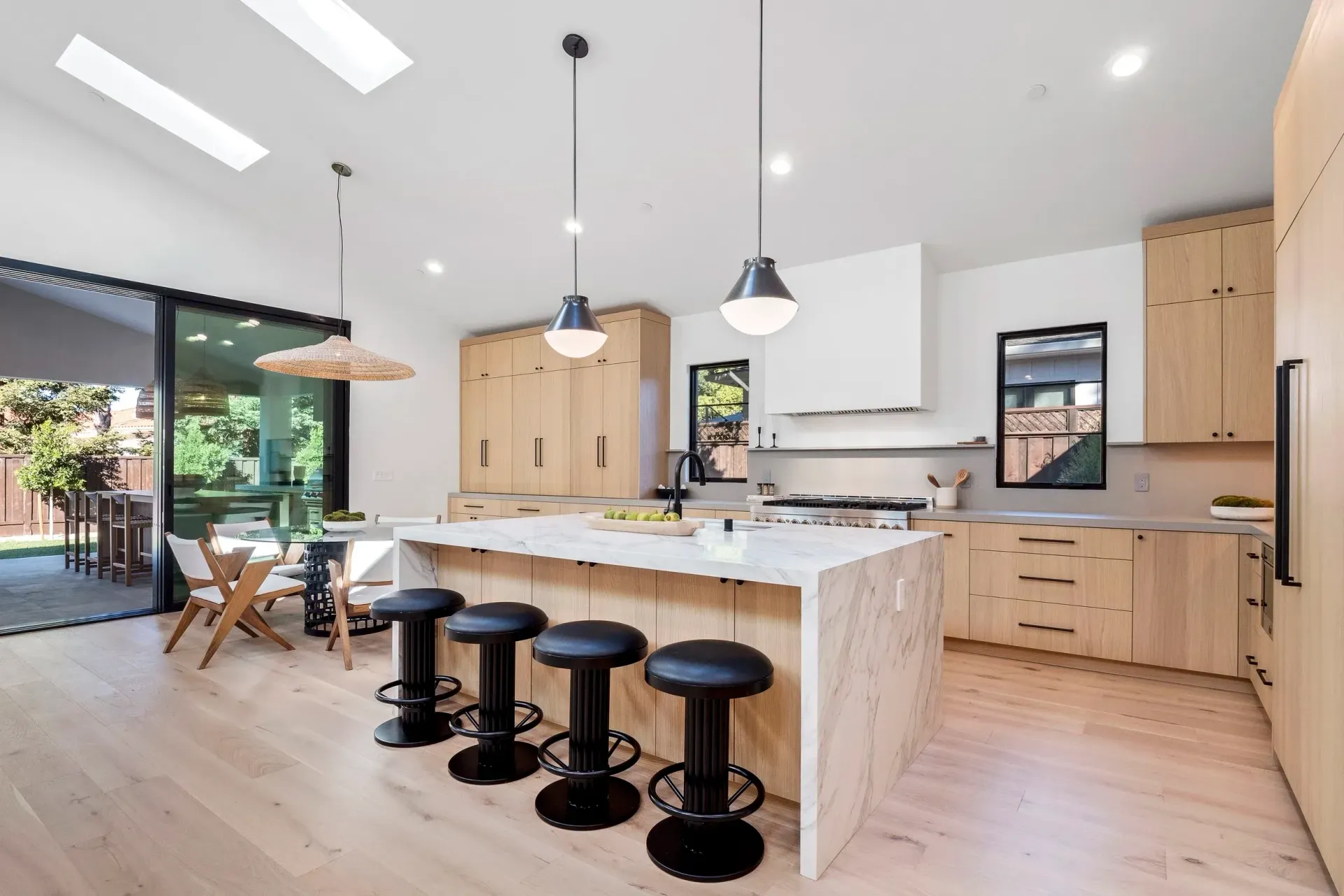 Modern kitchen with light wood cabinets, island with black stools, and outdoor access.