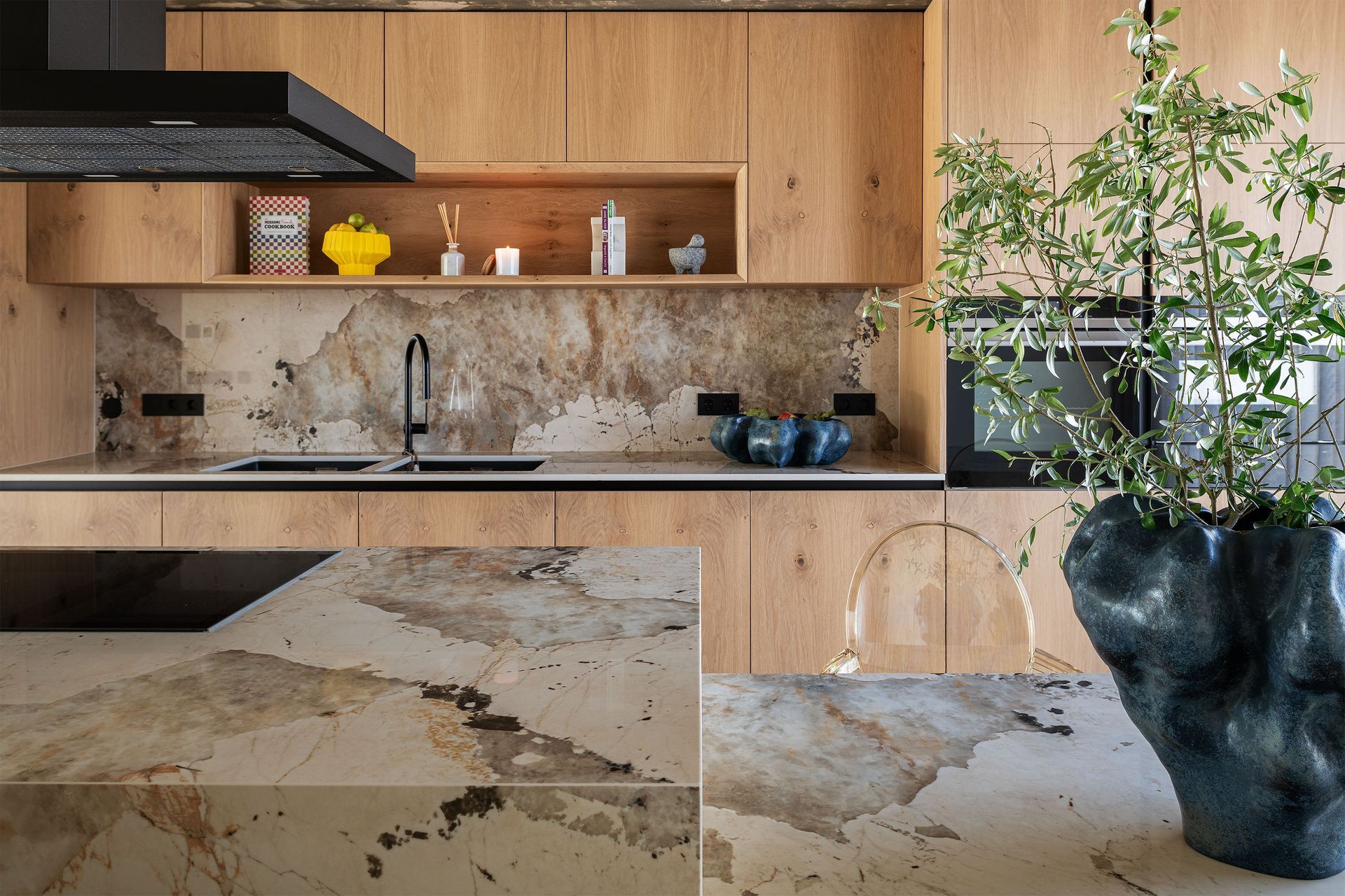 Modern kitchen with wood cabinets, marble backsplash, and a large plant in a dark vase.
