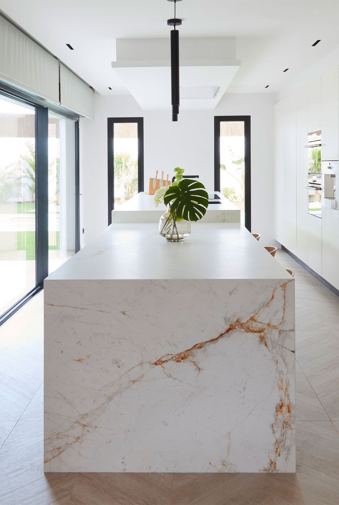 Modern white kitchen with large island, marble countertop with gold veining, and large leaf in vase.
