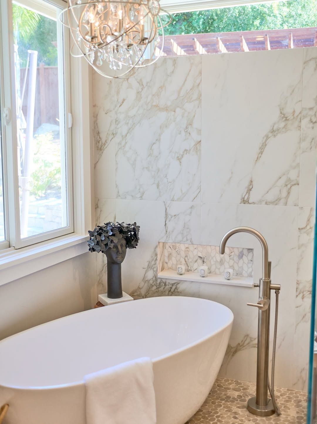 Bathroom with white oval tub, marble tile wall, silver faucet, and a window.