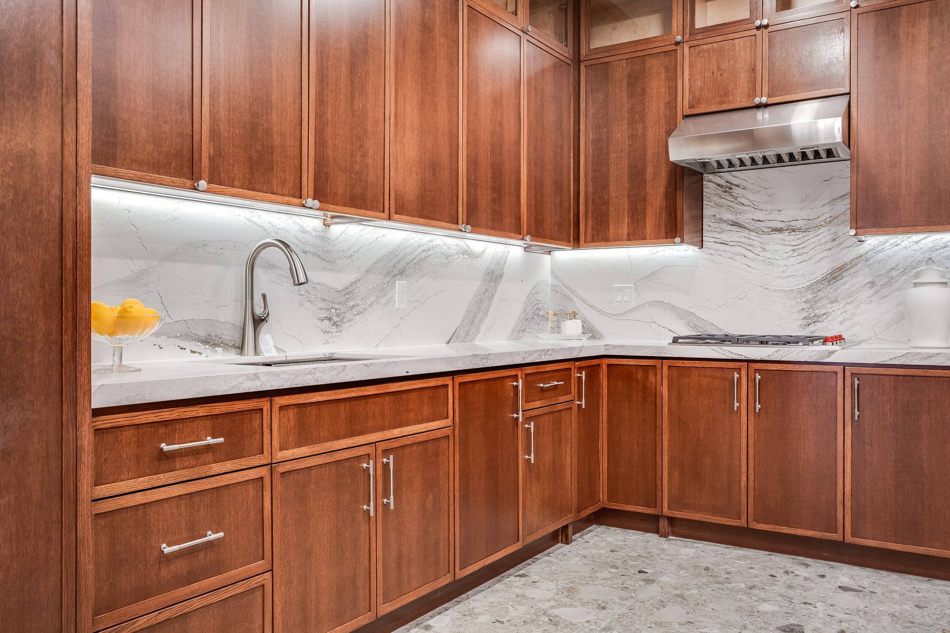 Wooden kitchen cabinets with marble backsplash, stainless steel range hood, and sink.