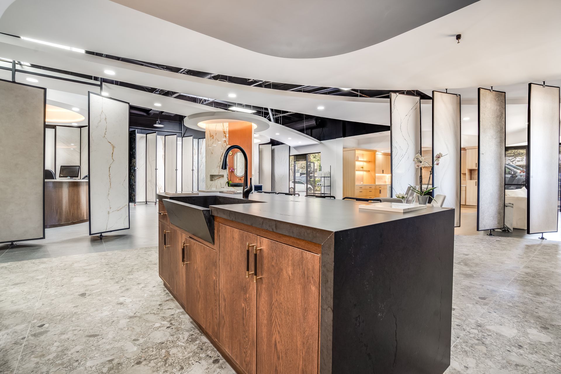 Reception desk in showroom. Curved brown base, light-colored top, computer, and two chairs under a circular recessed ceiling with lights.