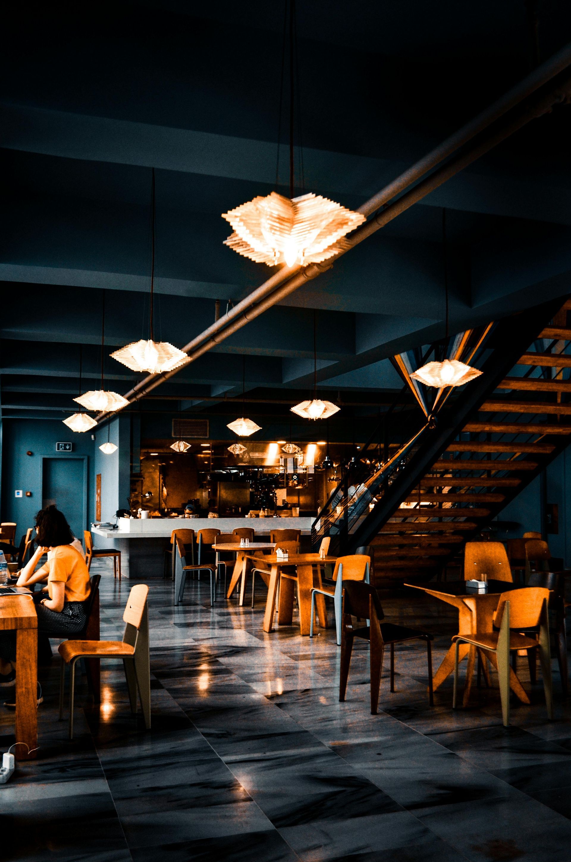 Restaurant interior with tables, chairs, and a bar area. Warm lights hang from the ceiling, reflecting on the floor.