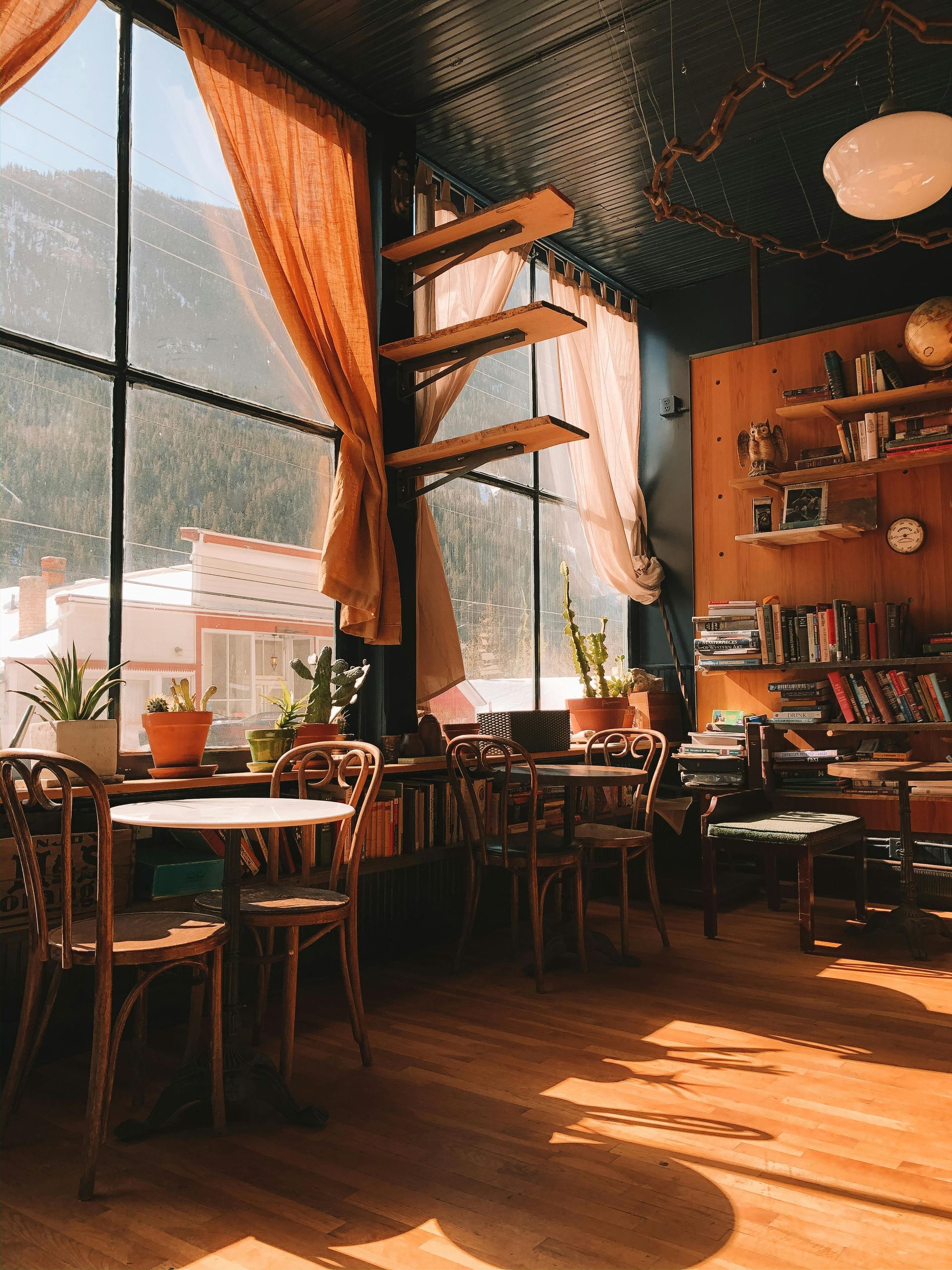 Cozy cafe interior with large window overlooking snowy mountain. Wooden tables, chairs, bookshelves.