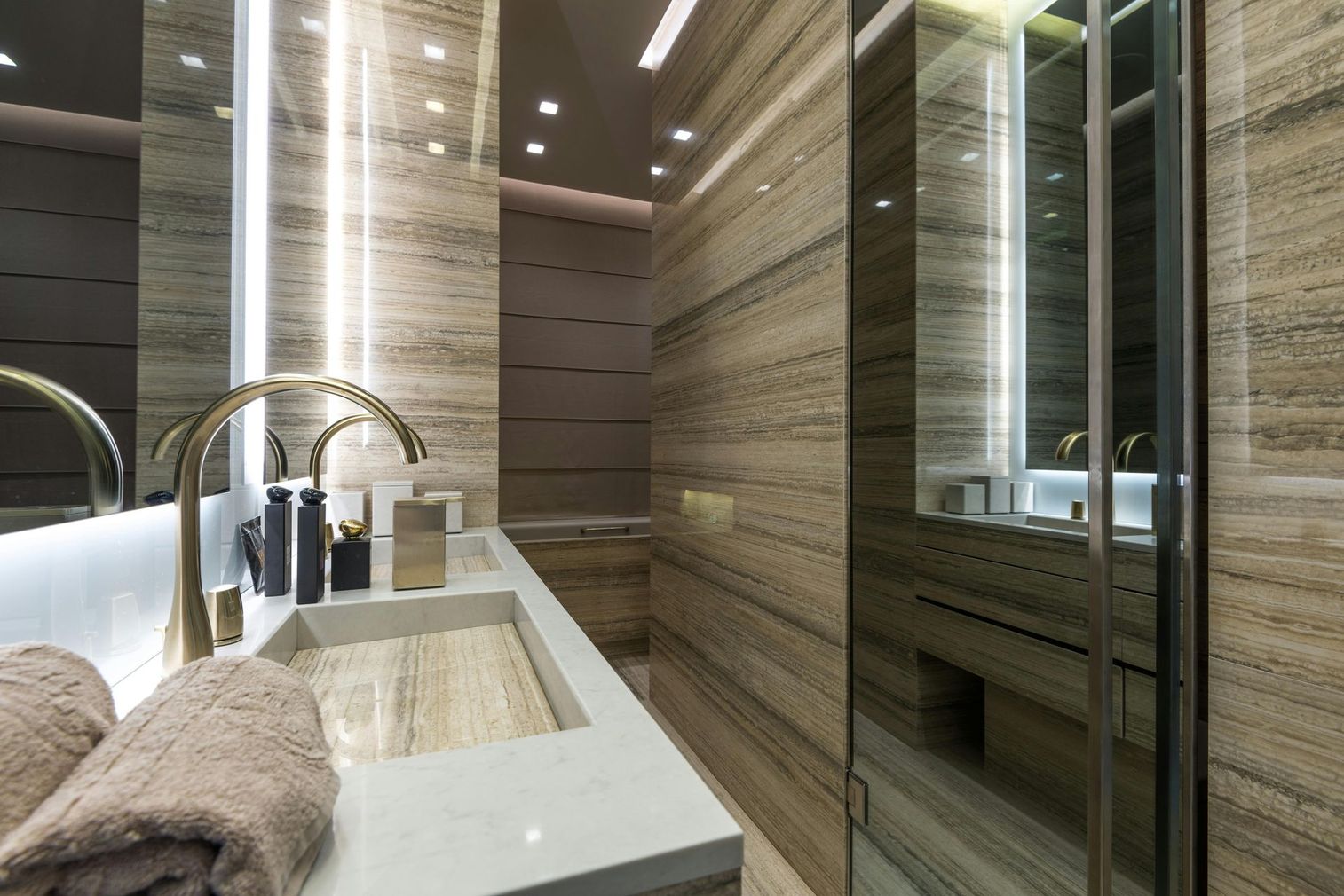 Modern bathroom with beige stone walls, gold fixtures, and long vanity with towels.