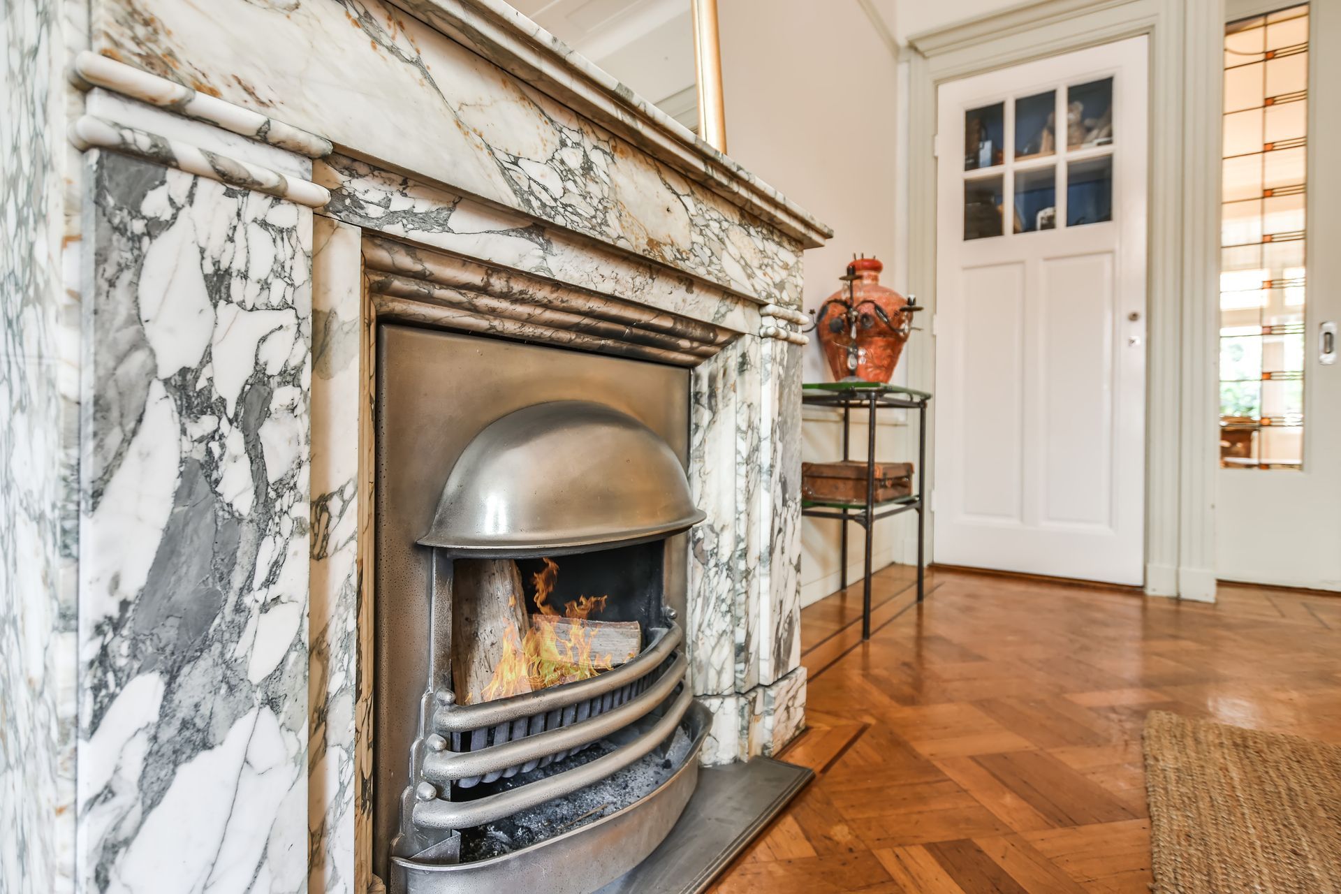 Marble fireplace with flames, beside a door and side table, on a parquet floor.