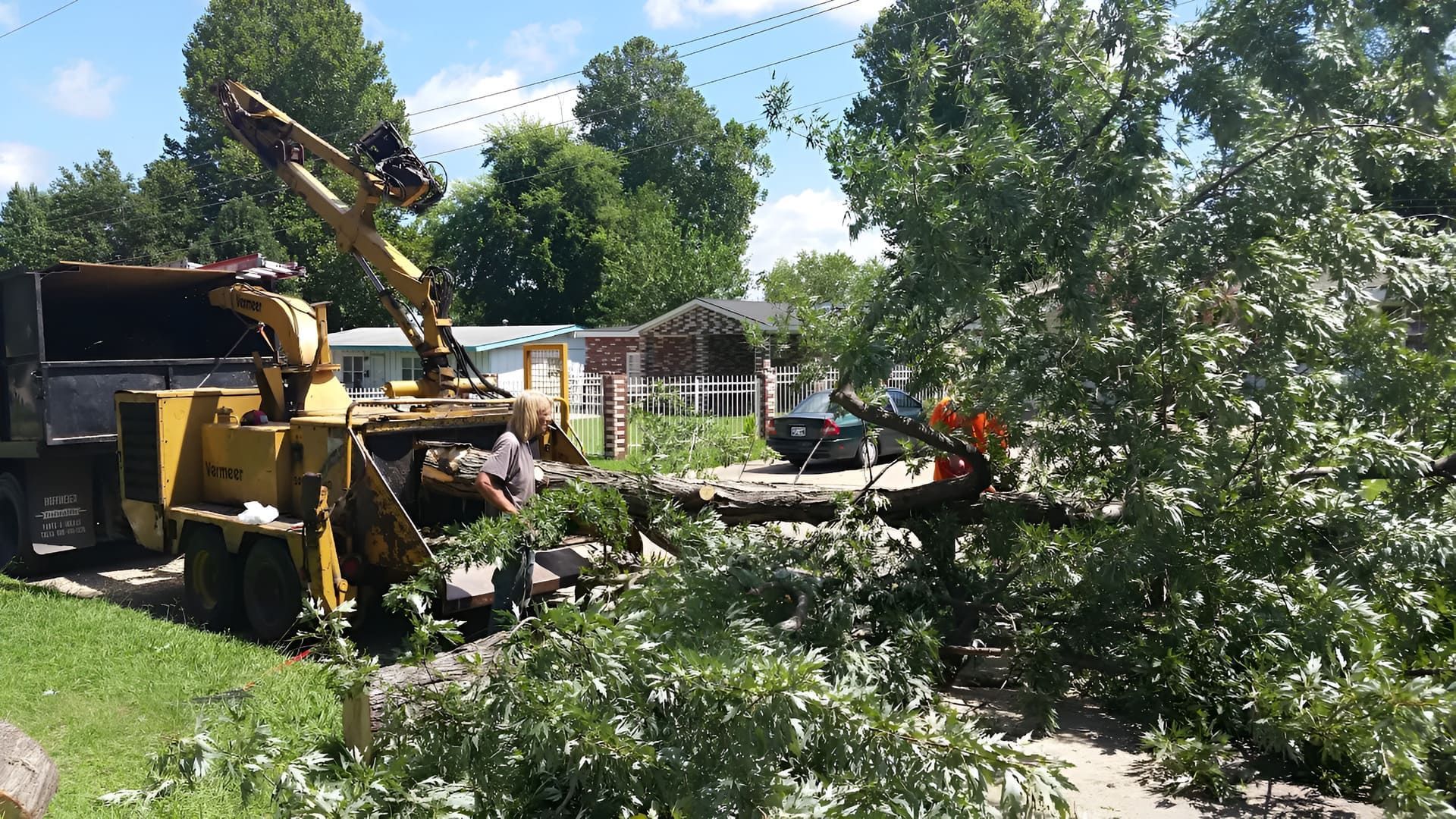A worker feeds a fallen tree into a yellow wood chipper parked on a residential street.