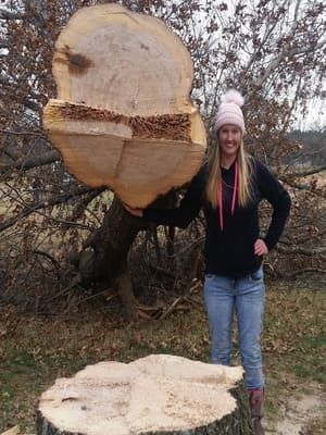 A smiling person standing beside a large, freshly cut tree stump, holding up a matching cross-section of the trunk.
